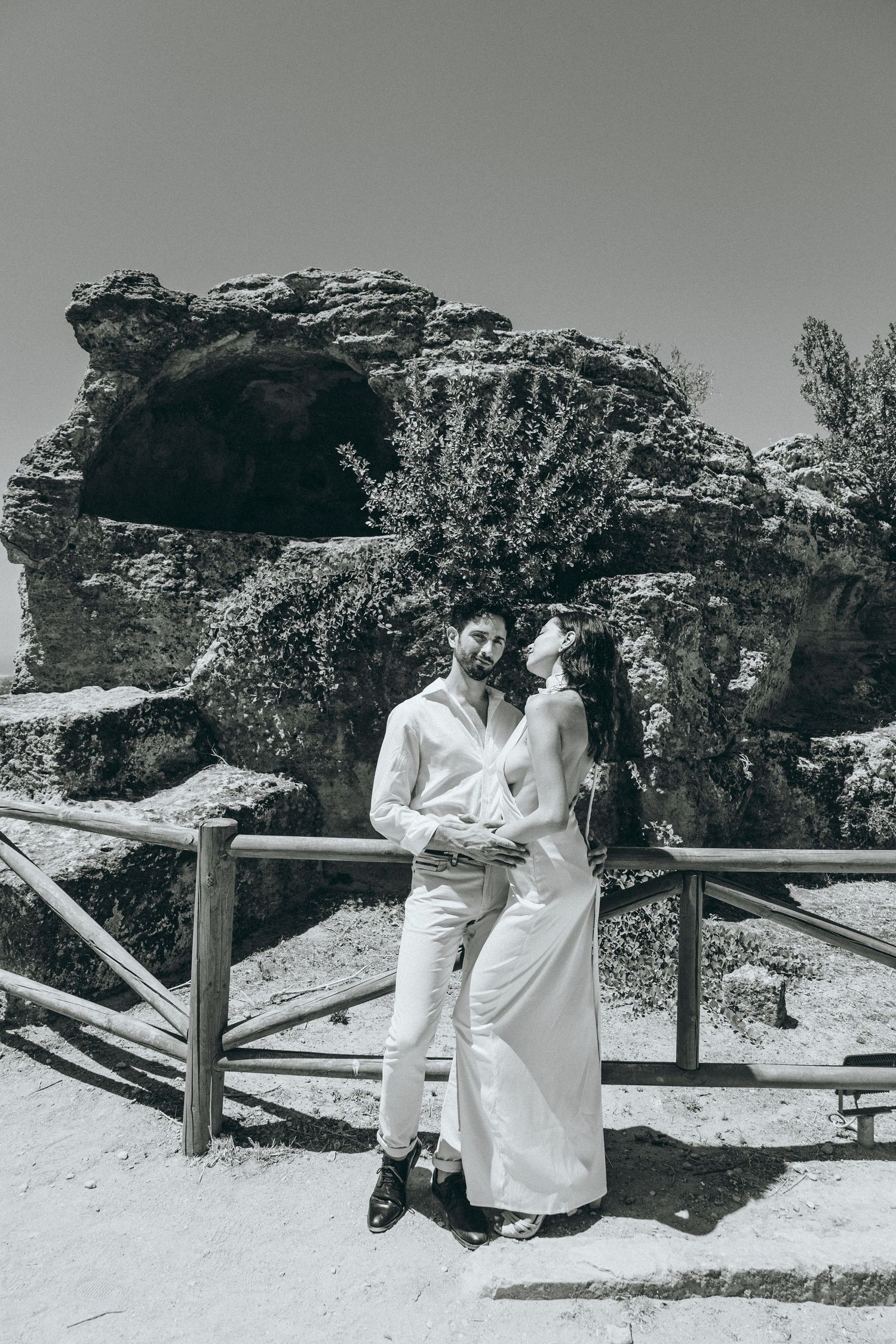 Couple shares tender moments along rustic wooden fence with rocky landscape behind.