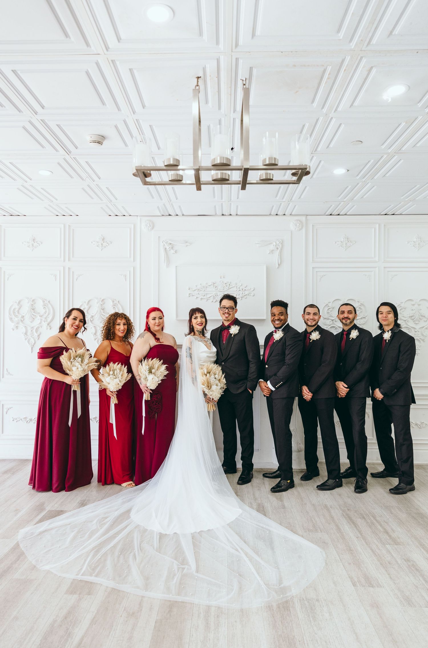 Full wedding party in burgundy and black attire poses elegantly in a white room with chandelier.