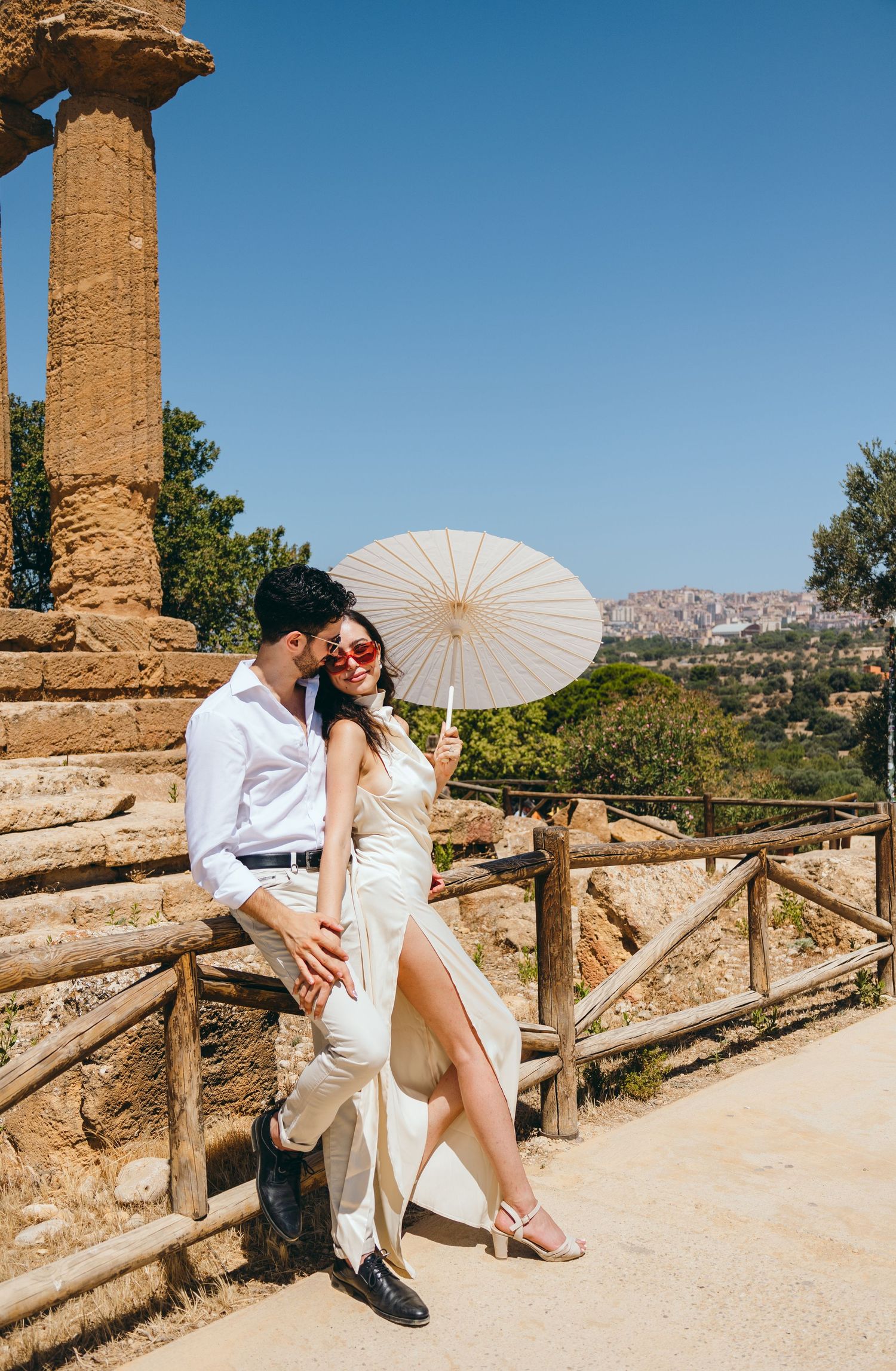 Couple posing with white parasol at ancient ruins in Sicily with scenic mountain landscape in background.