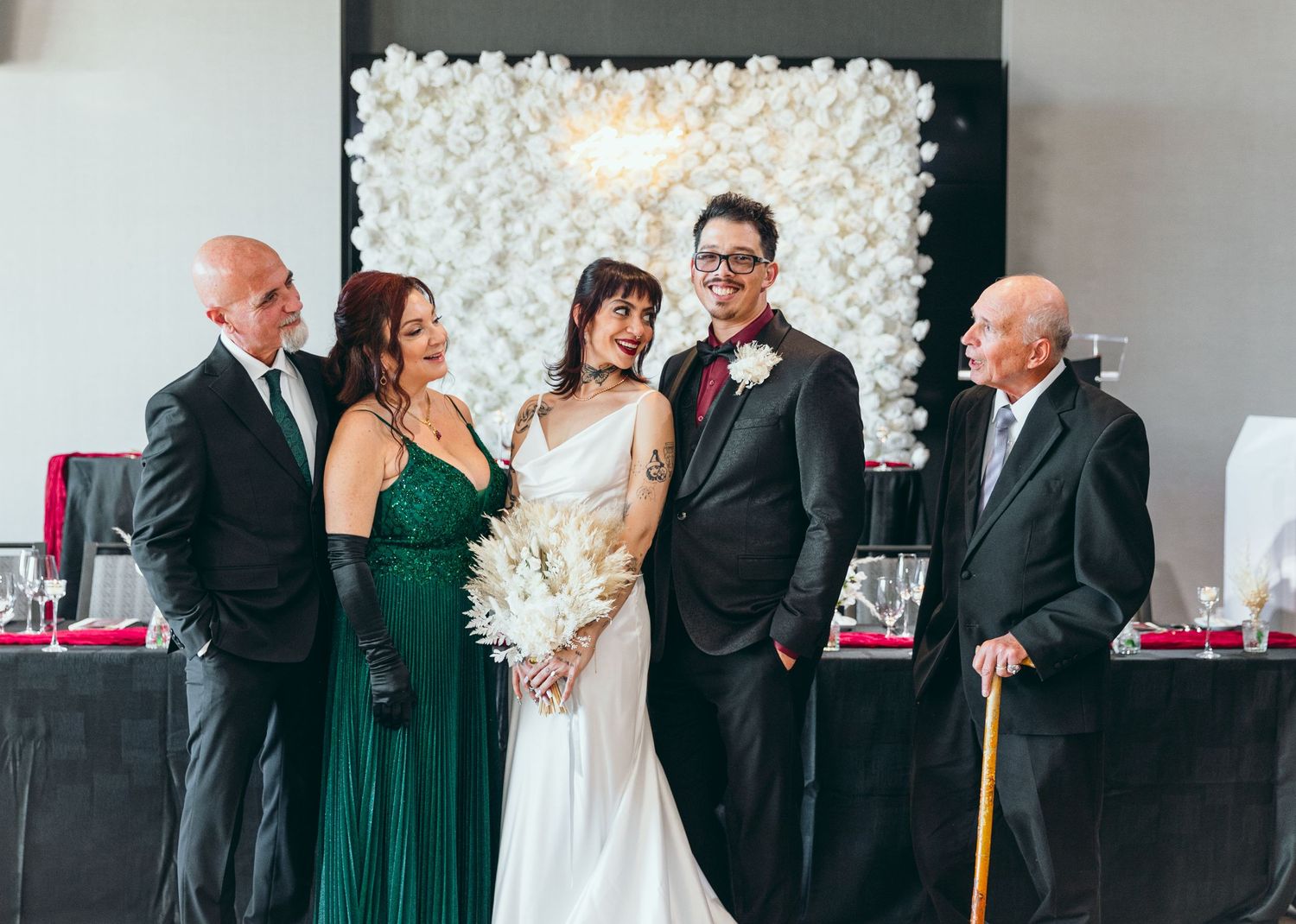Wedding party gathers for photos in front of white floral wall with elegant decor.