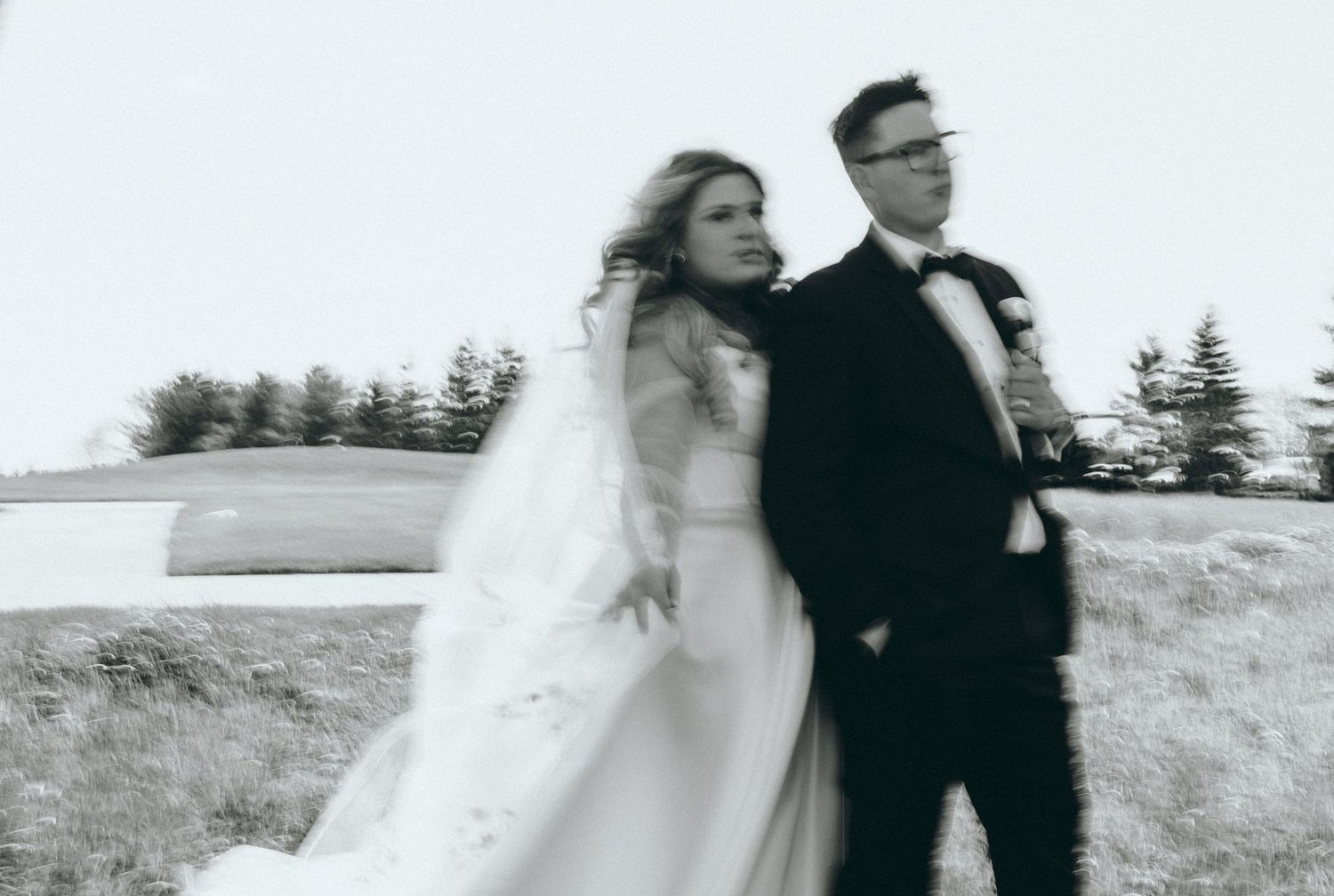 Black and white motion-blurred portrait of newlyweds outdoors with trees in background.