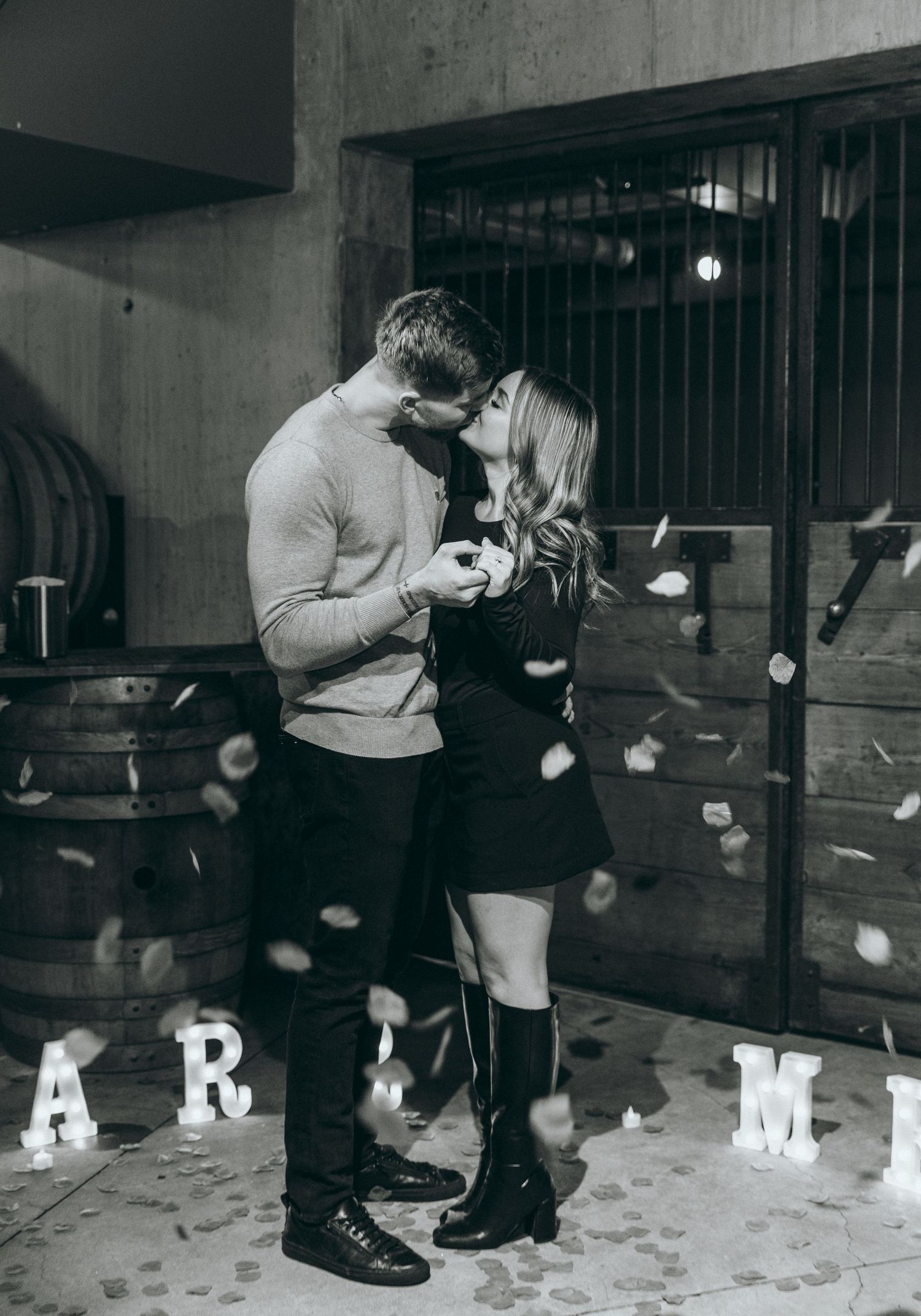 A romantic proposal moment in a cellar with scattered rose petals and illuminated letters spelling out 'MARRY ME'.