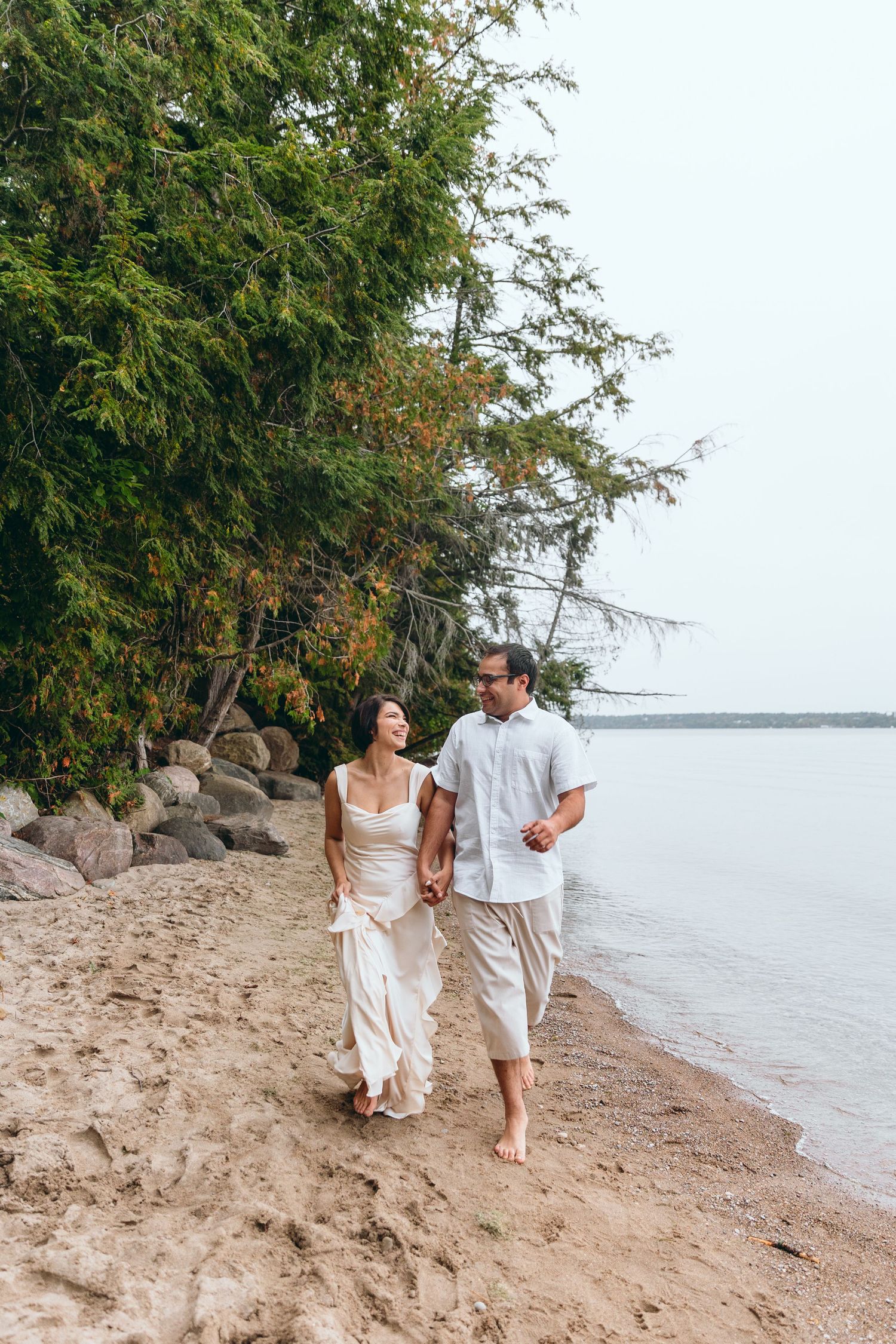 A couple in white outfits walks along a sandy beach shoreline.