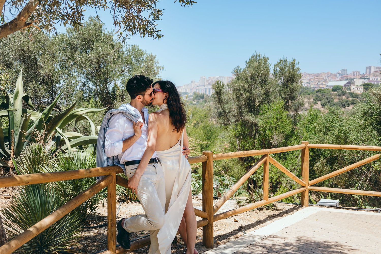 A romantic moment captured on a path with wooden railings and olive trees in the background.