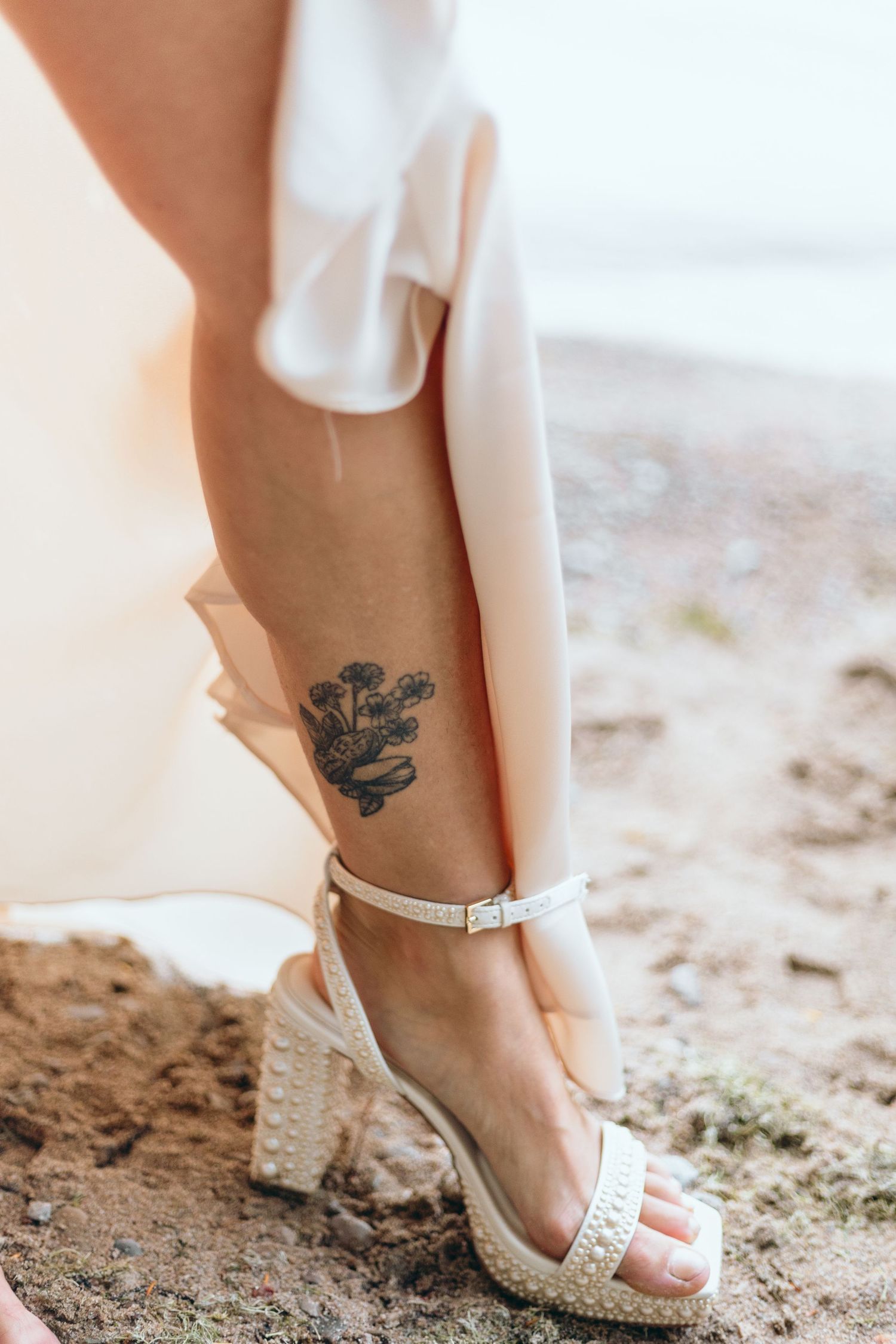 White heeled sandal and small oak leaf tattoo on ankle against sandy beach background.