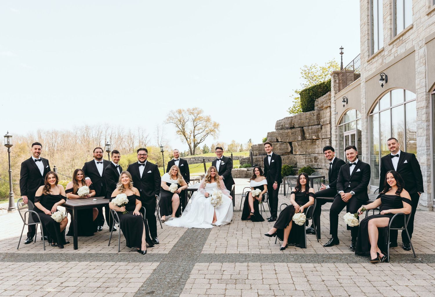 Wedding party sits casually on stone steps during outdoor celebration.