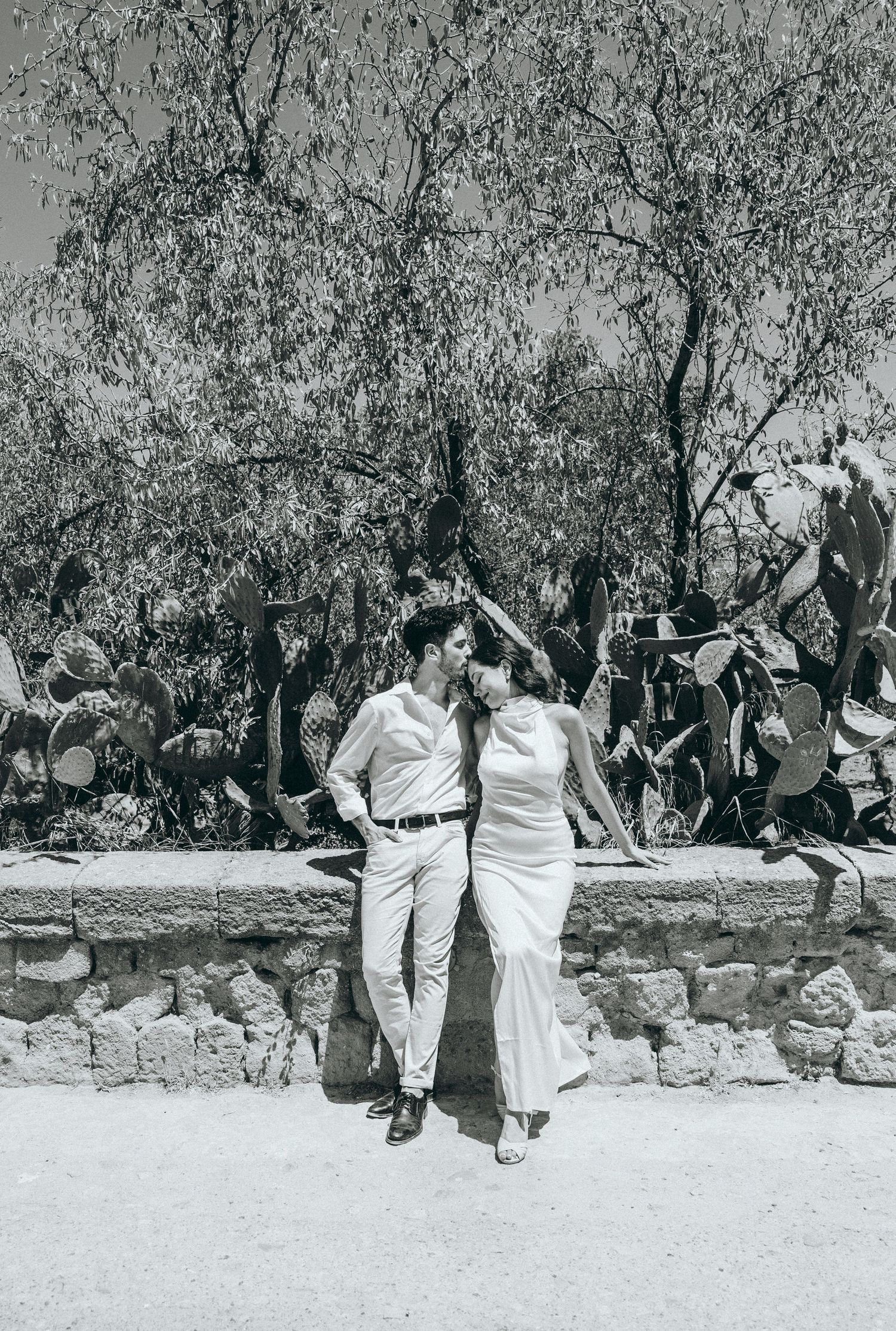 A couple in white outfits poses playfully on a stone wall with trees in the background.