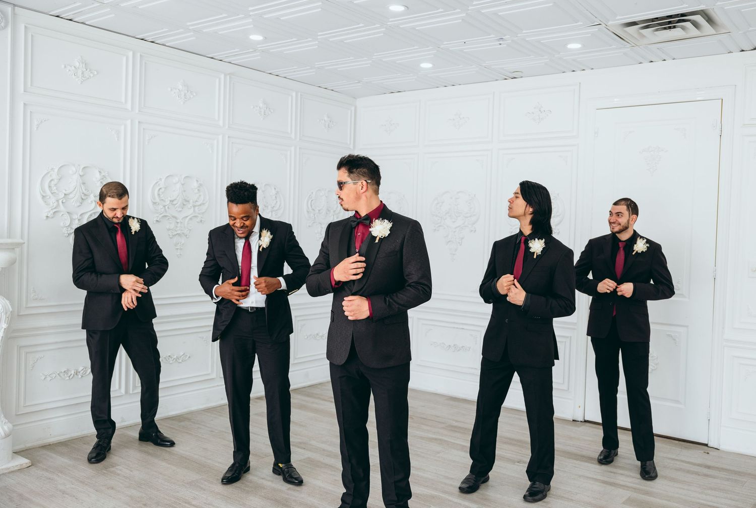 Groomsmen in black suits pose in line against white wall.