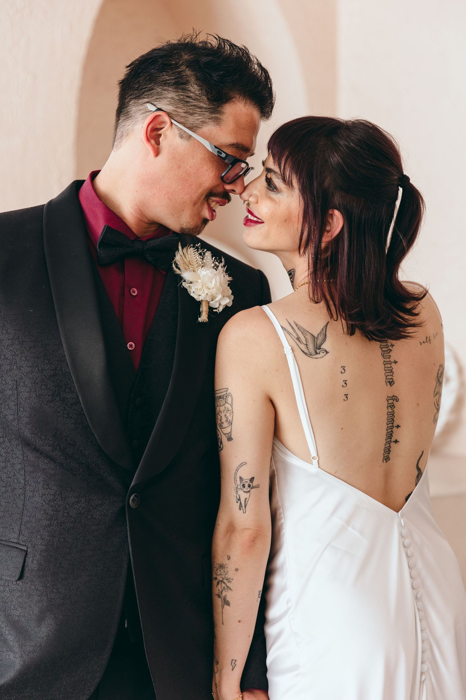 An intimate wedding portrait showing tattoos and formal attire against a light background.