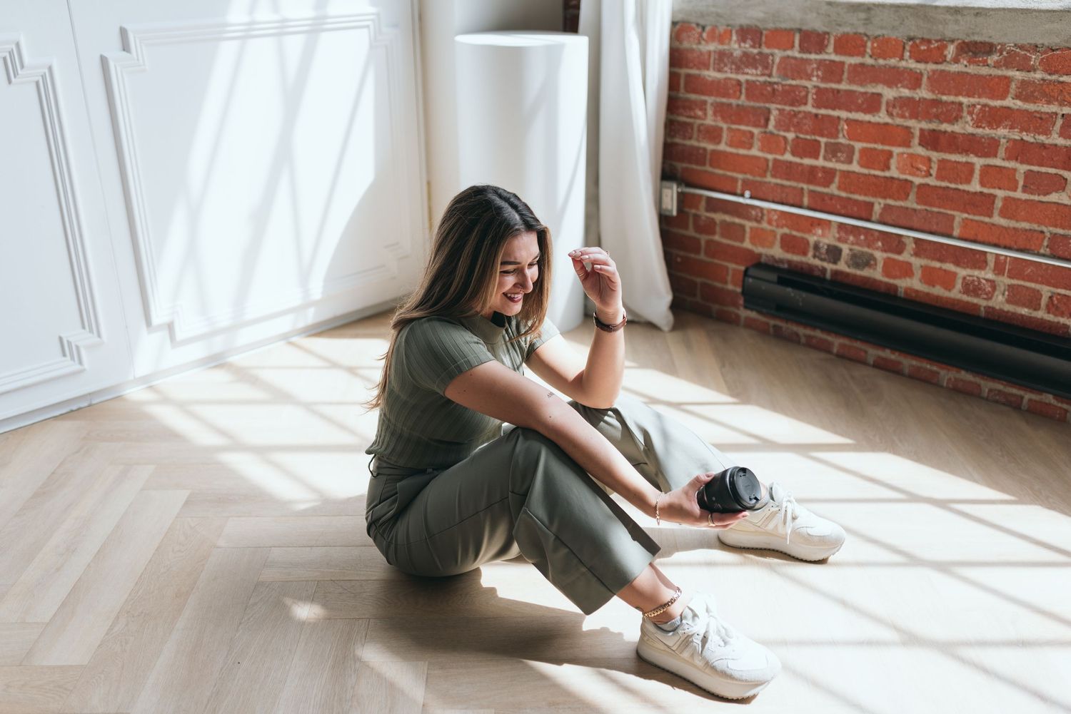 Woman sitting in sunlit room with hardwood floors and exposed brick wall.