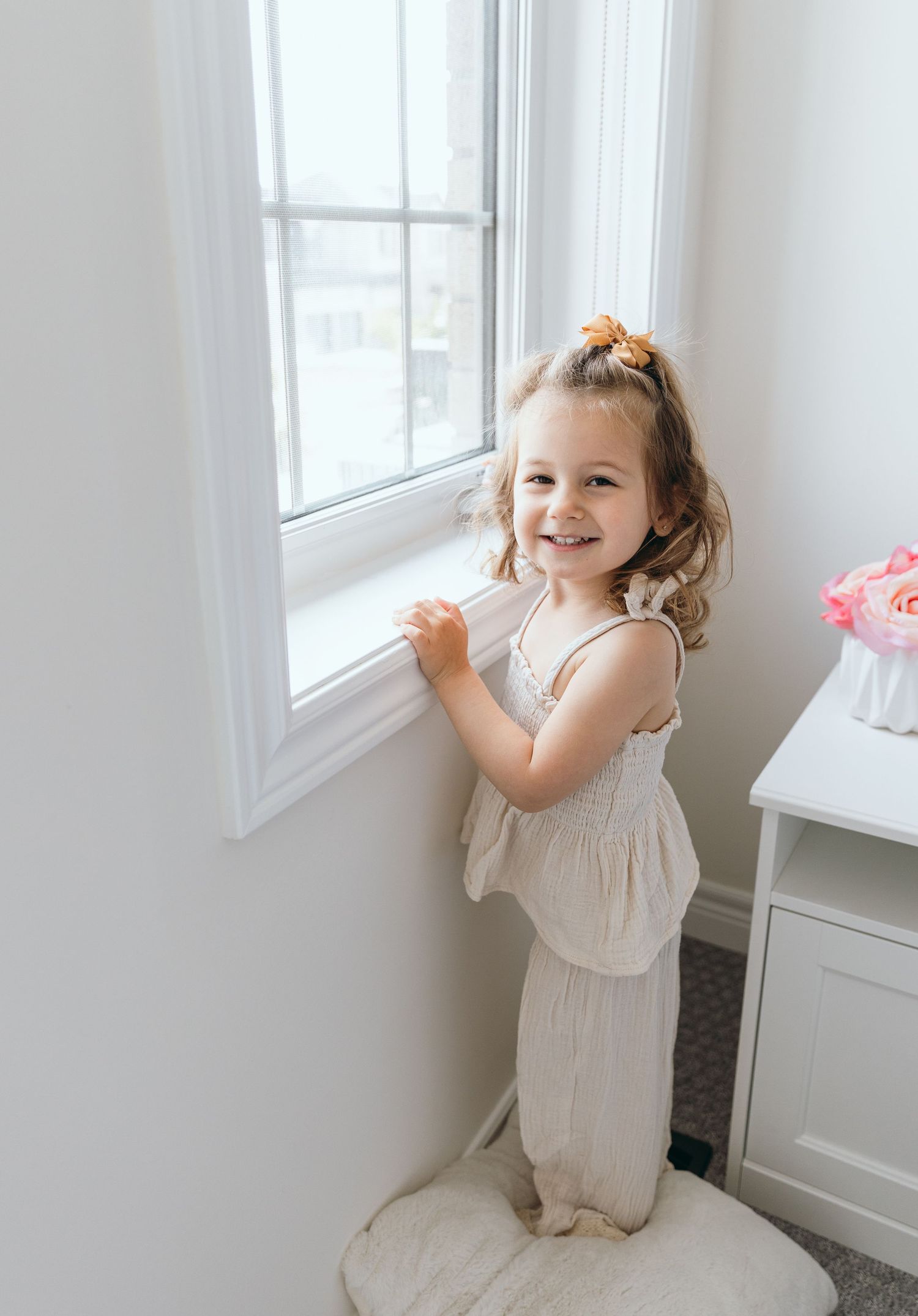 A toddler in a white dress plays and dances joyfully near a bright window.