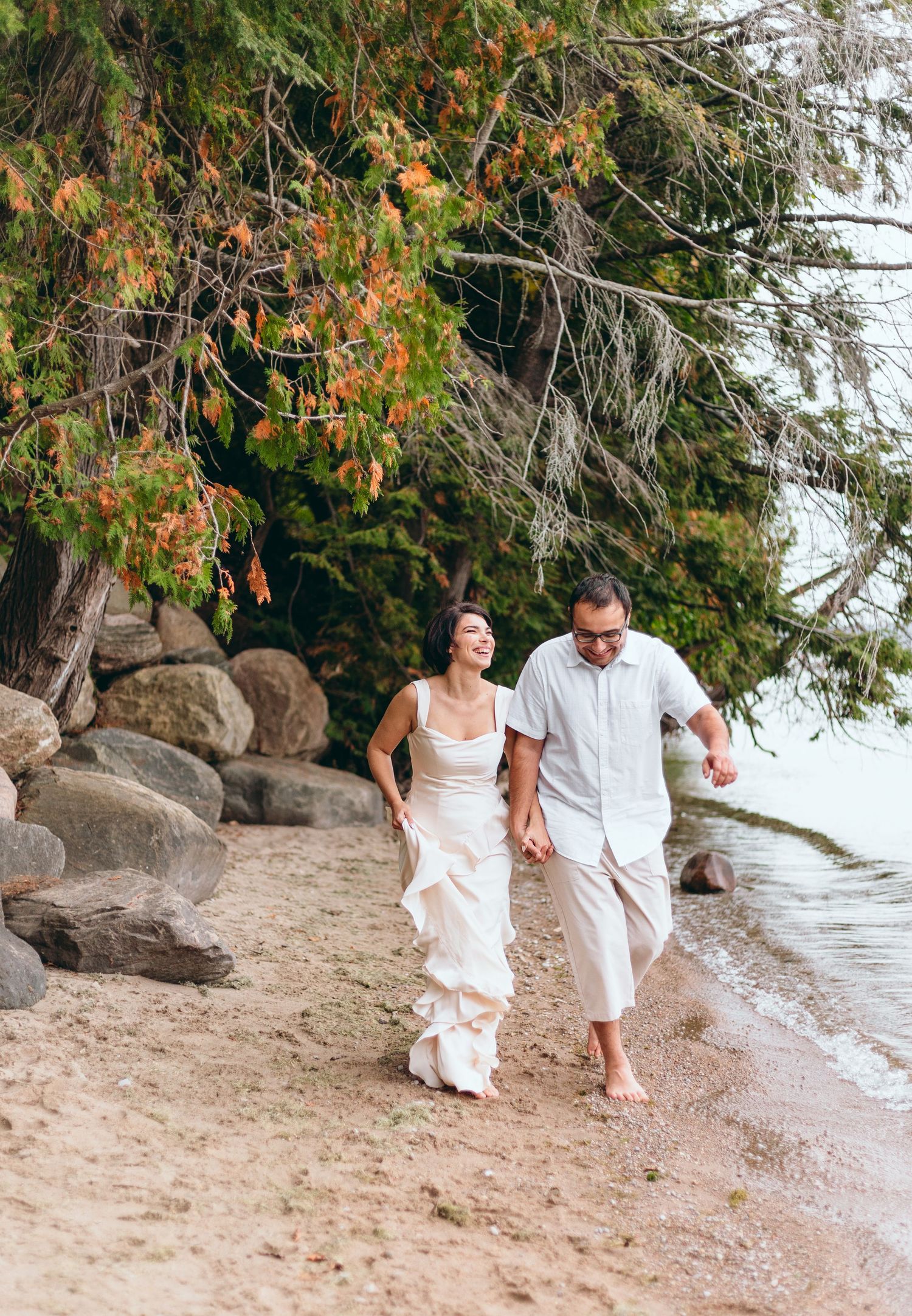 Couple walking hand in hand along a scenic beach path under evergreen trees.