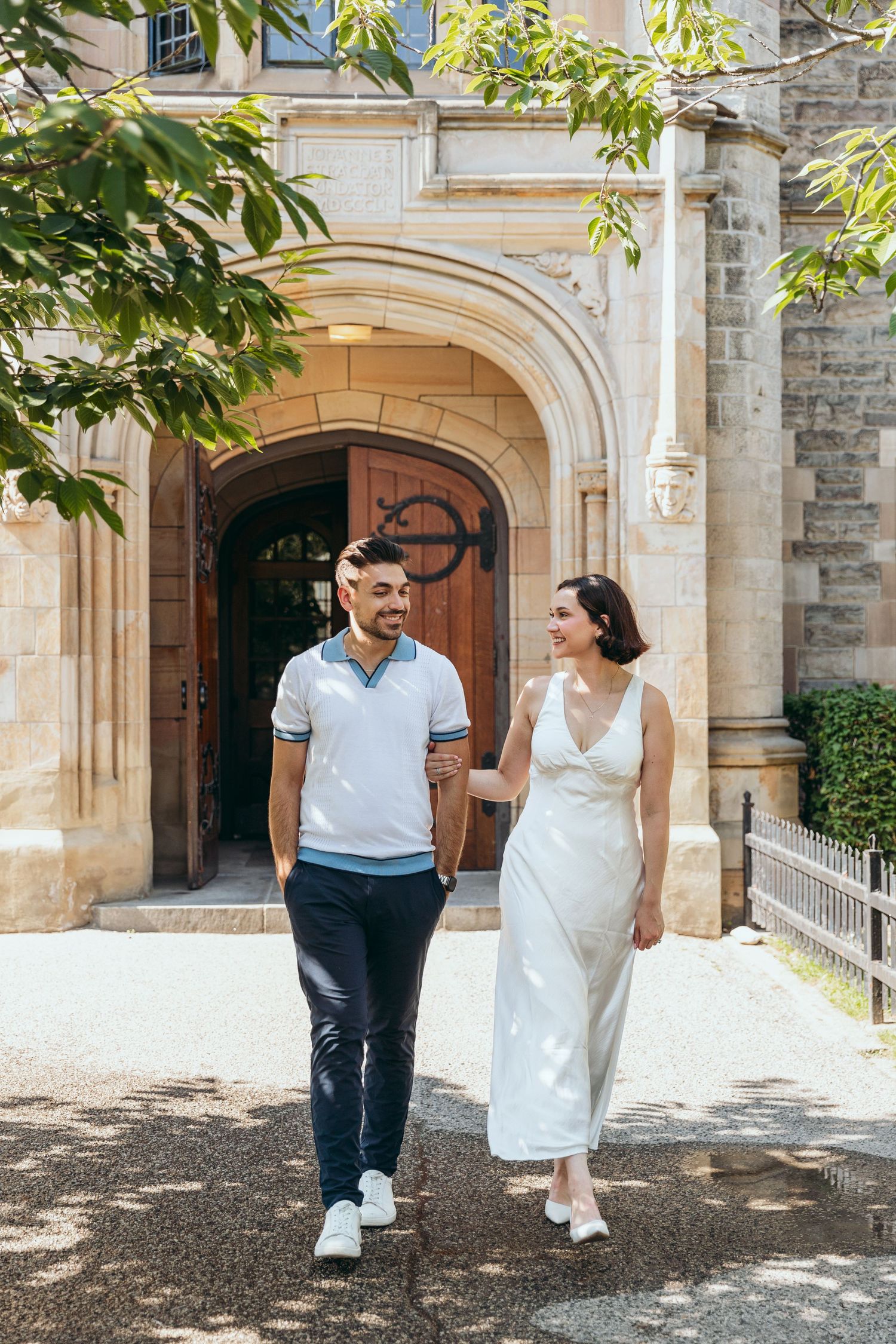 Couple walks hand in hand along a tree-lined path by a historic stone building.