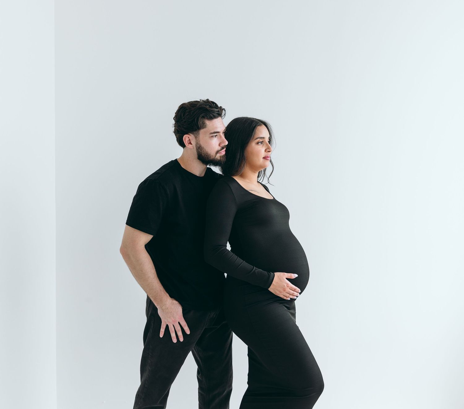 Maternity photos of couple in matching black outfits against white backdrop.