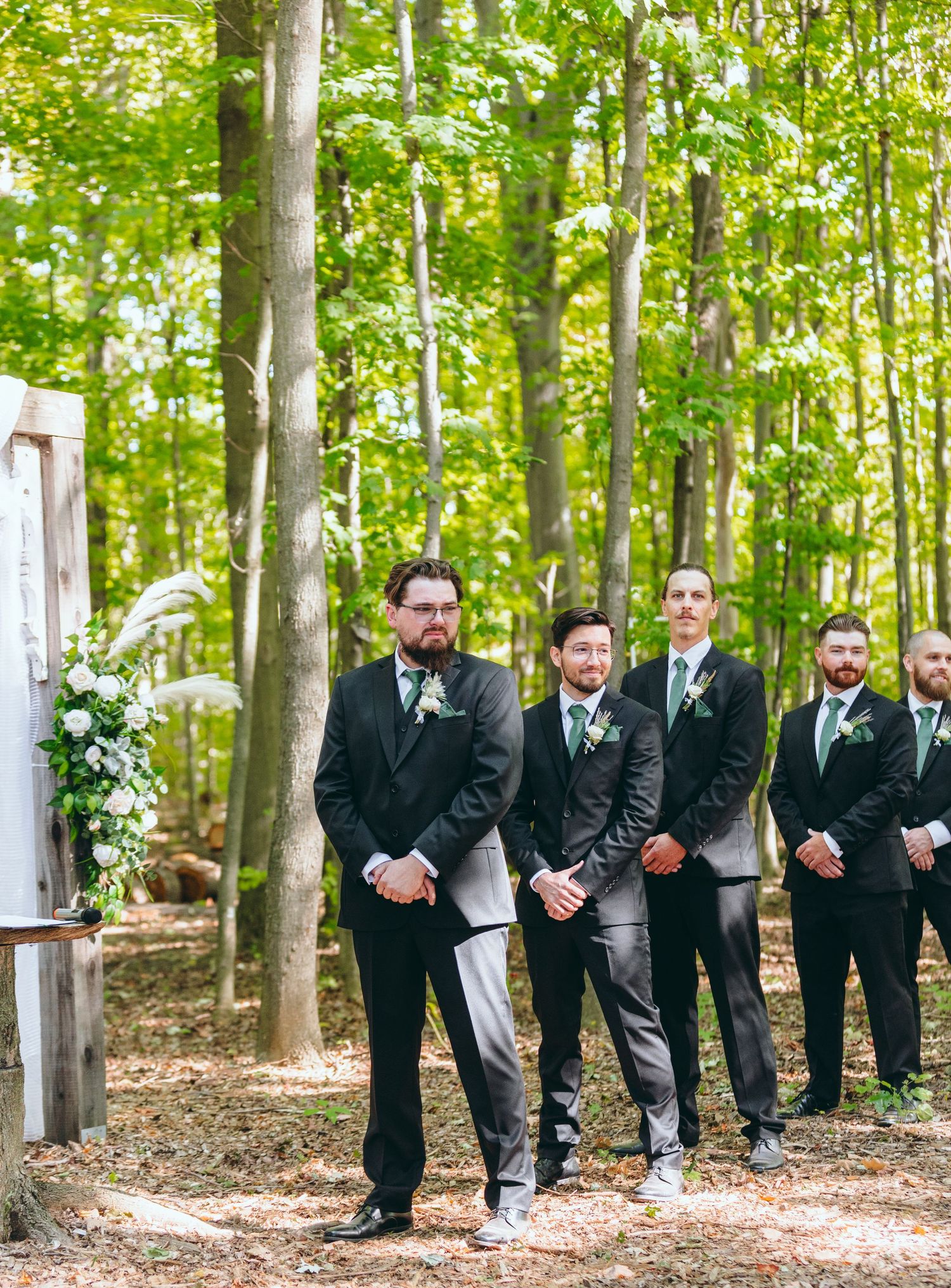 Groomsmen in black suits pose together in a wooded outdoor setting.