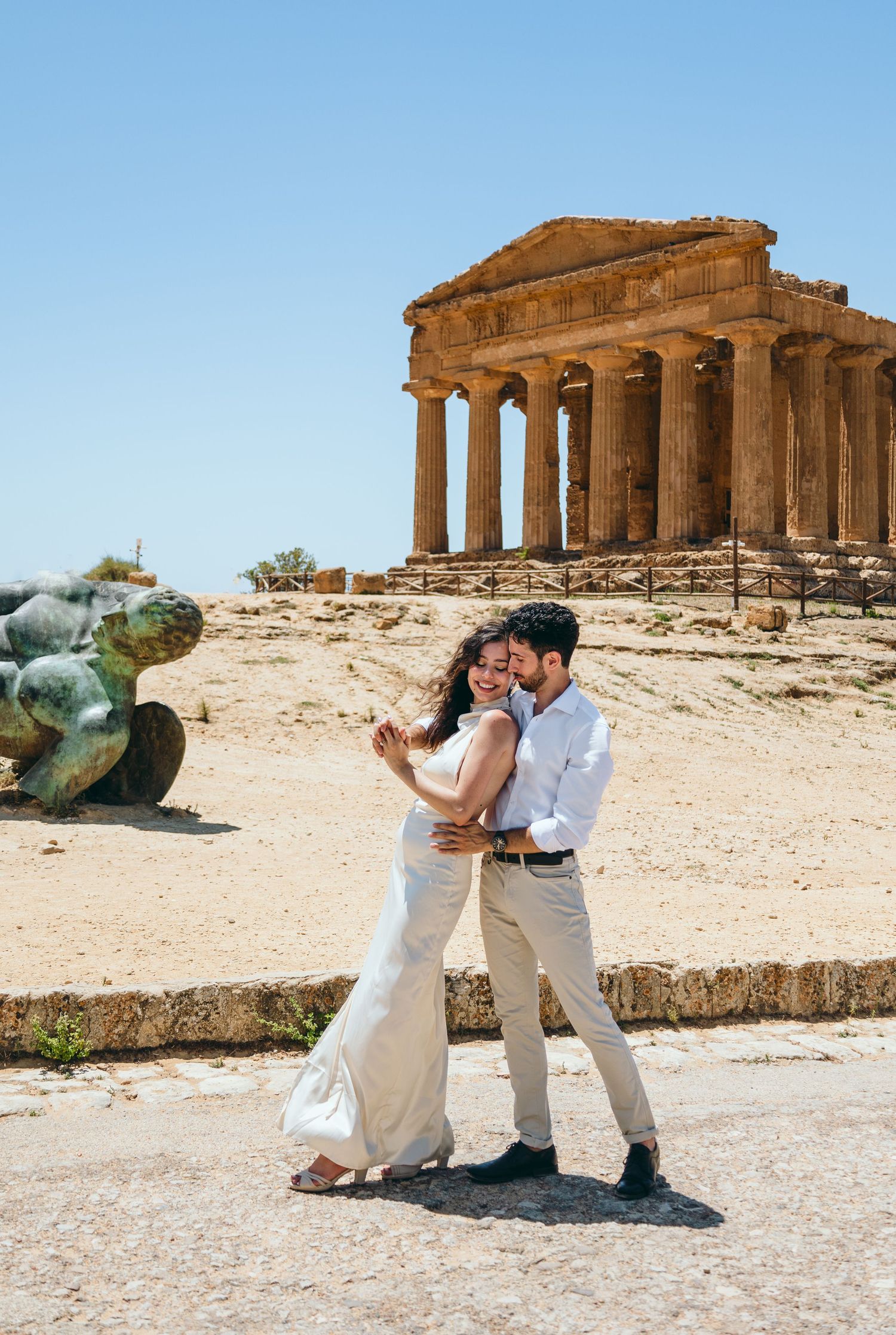 Couple shares intimate moment near fallen statue with Greek temple ruins in Sicily.