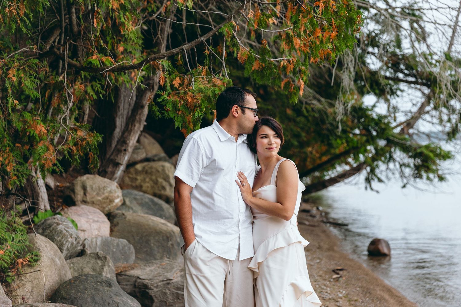 A couple in white outfits standing together on a rocky beach with trees overhead.