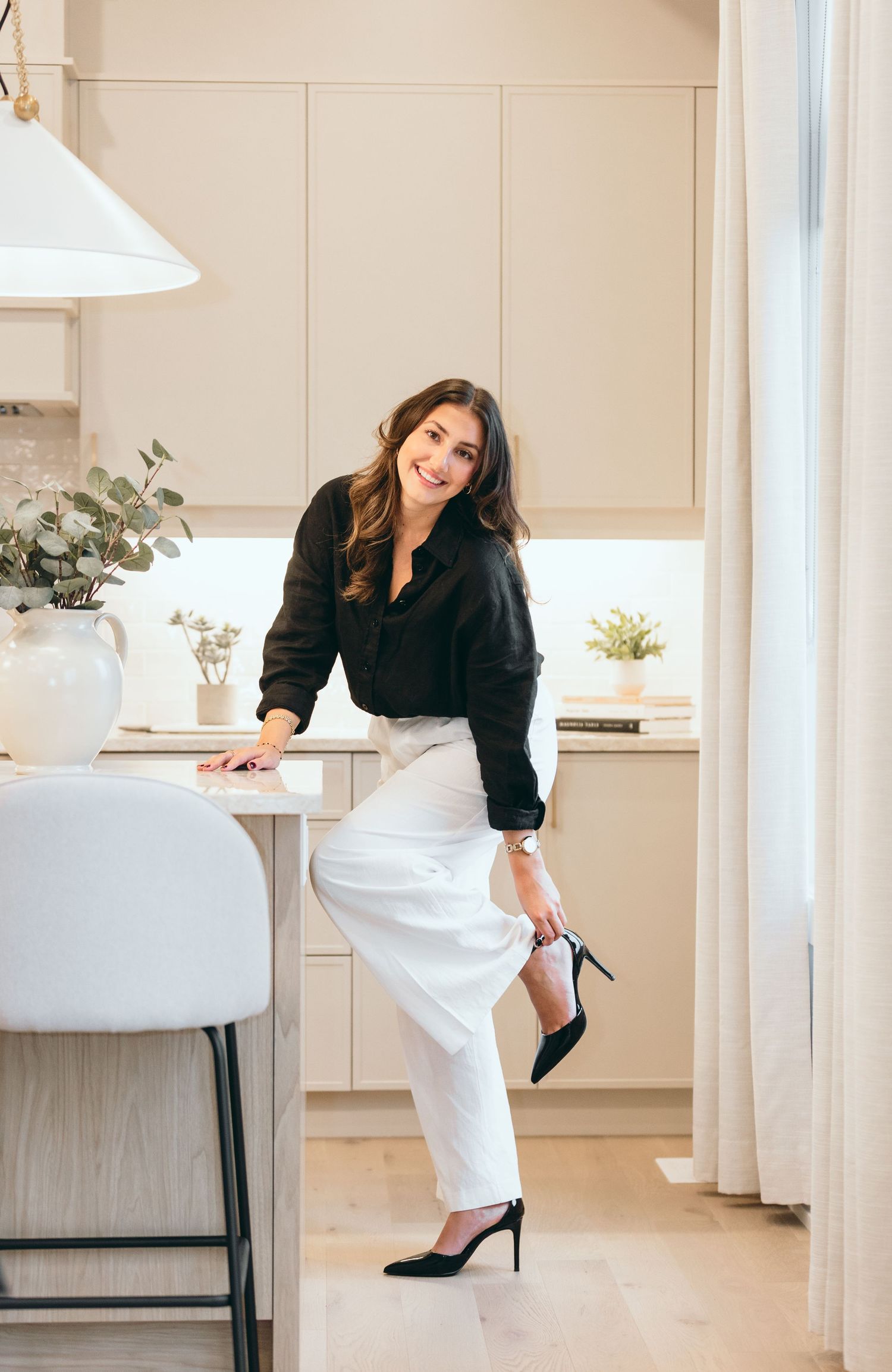 Stylish woman in black blazer and white pants poses elegantly in a modern kitchen setting.