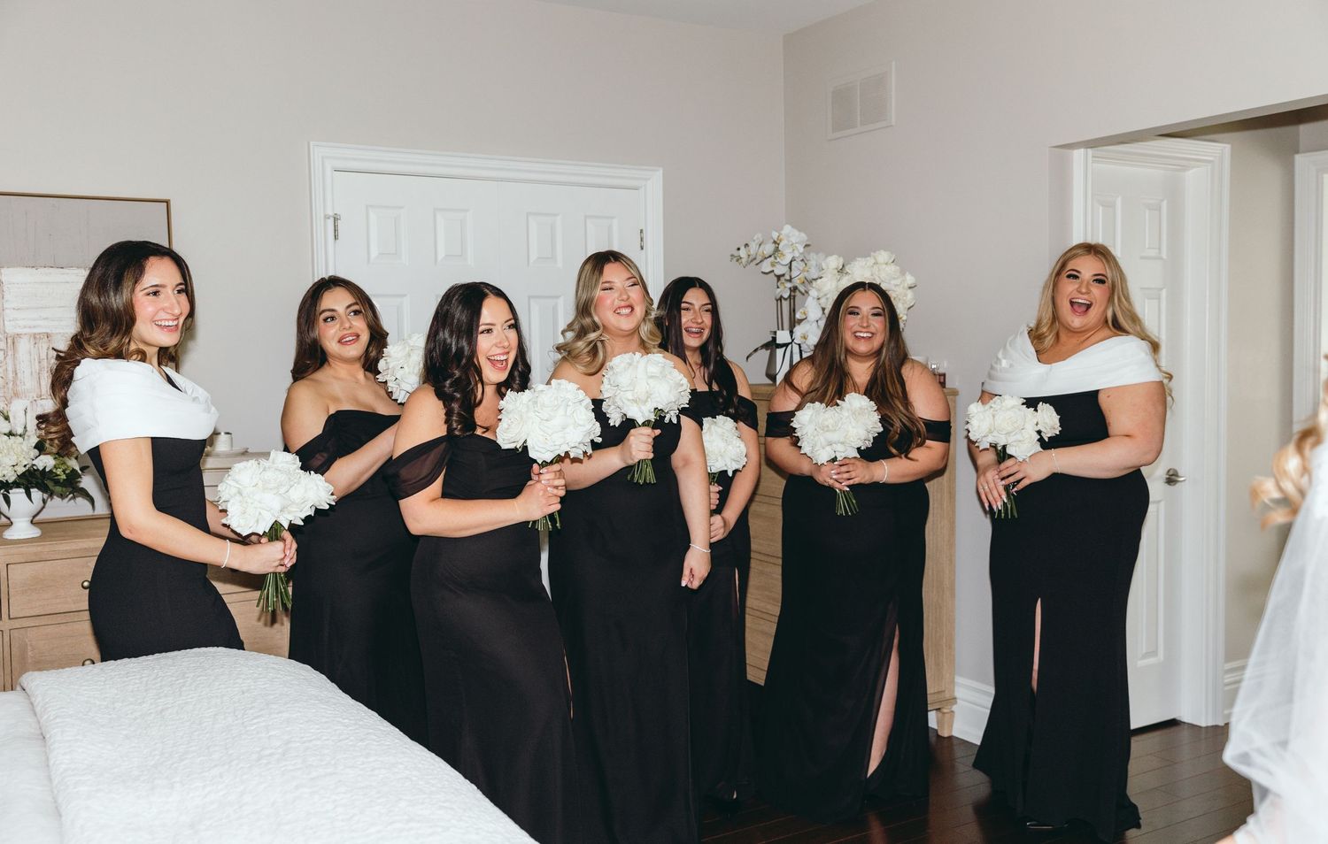 Wedding party in black dresses poses together in bright indoor setting.