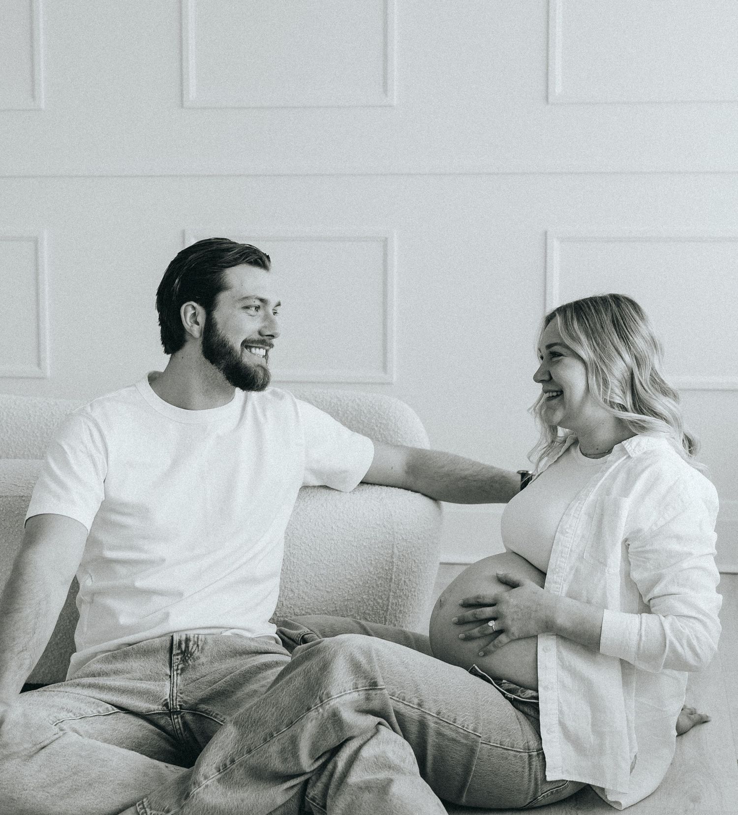 Couple relaxing on white couch during casual pregnancy photoshoot.
