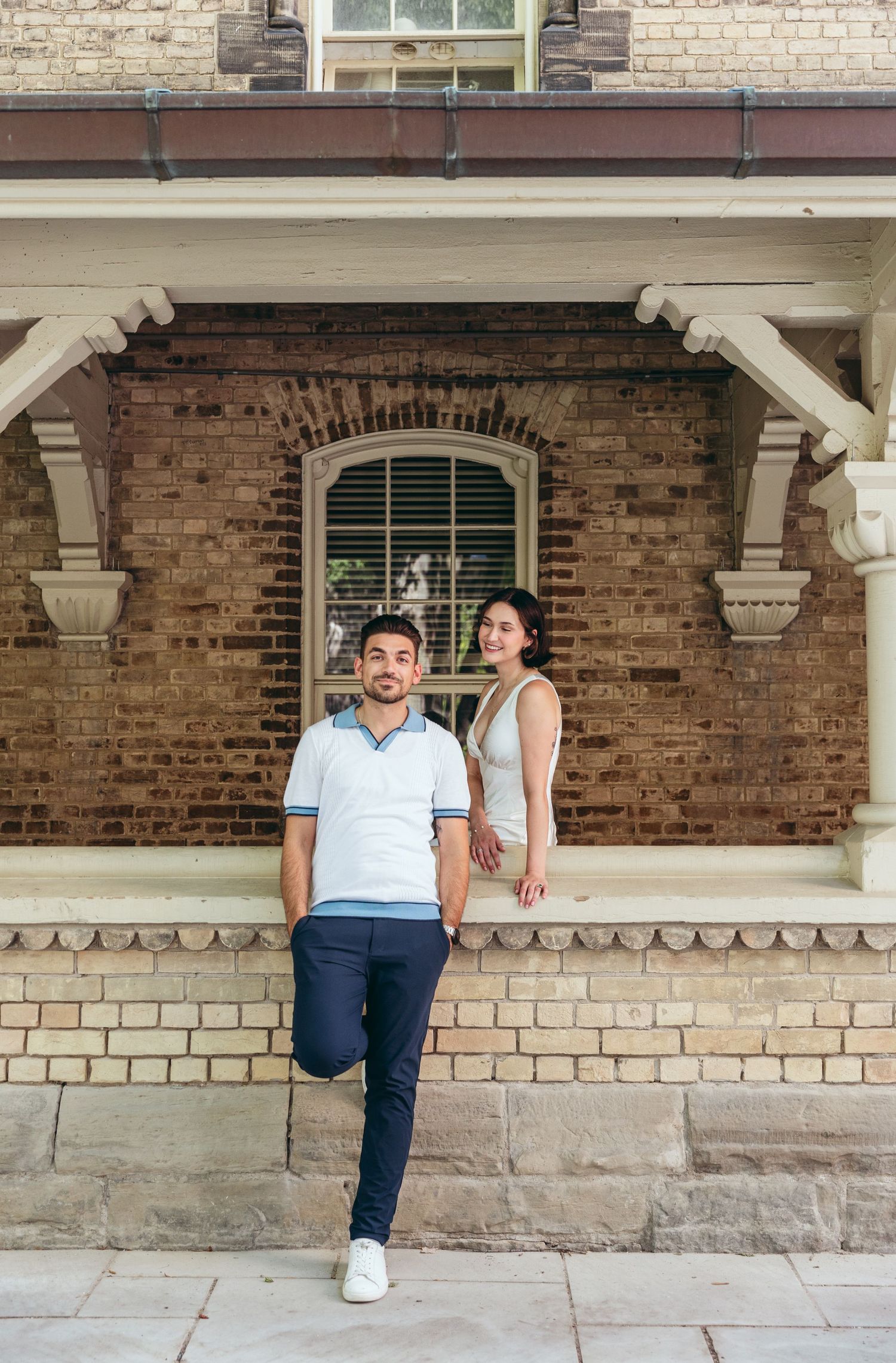 A couple strikes a fun pose against brick walls and decorative stone columns.