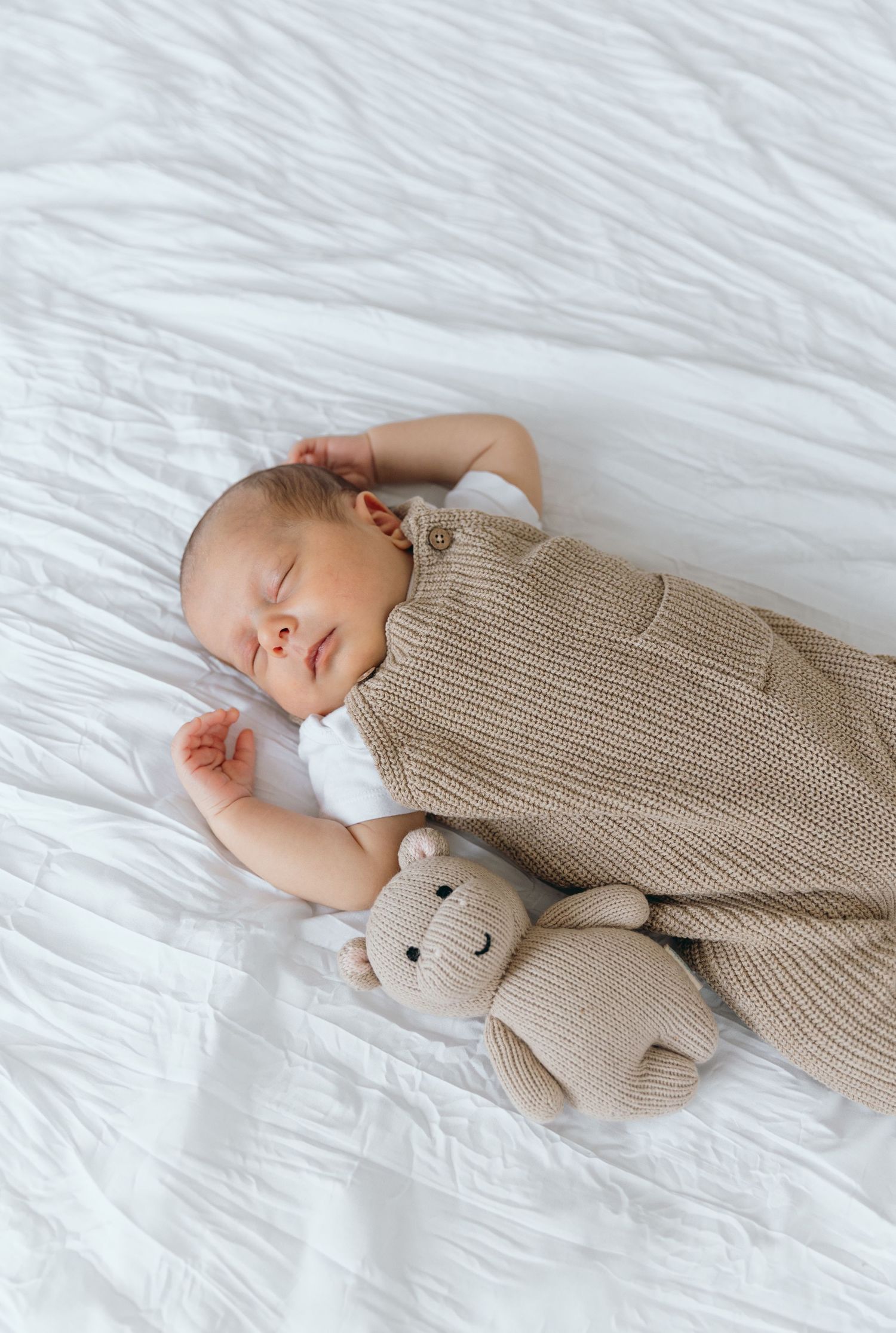 Baby resting peacefully on white sheets while cuddling soft beige teddy bear.