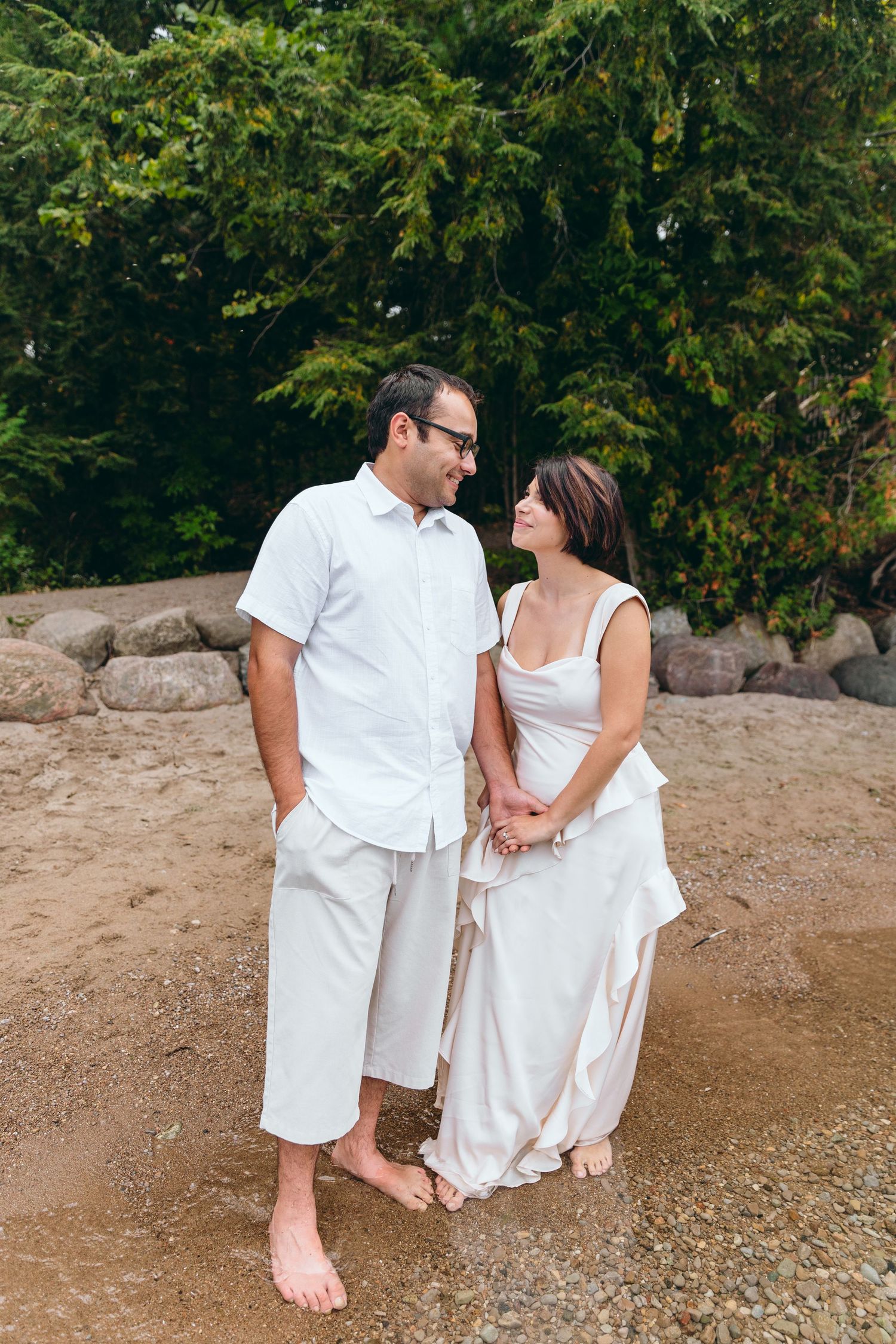 Romantic couple in white outfits sitting together on rocky beach surrounded by green trees.