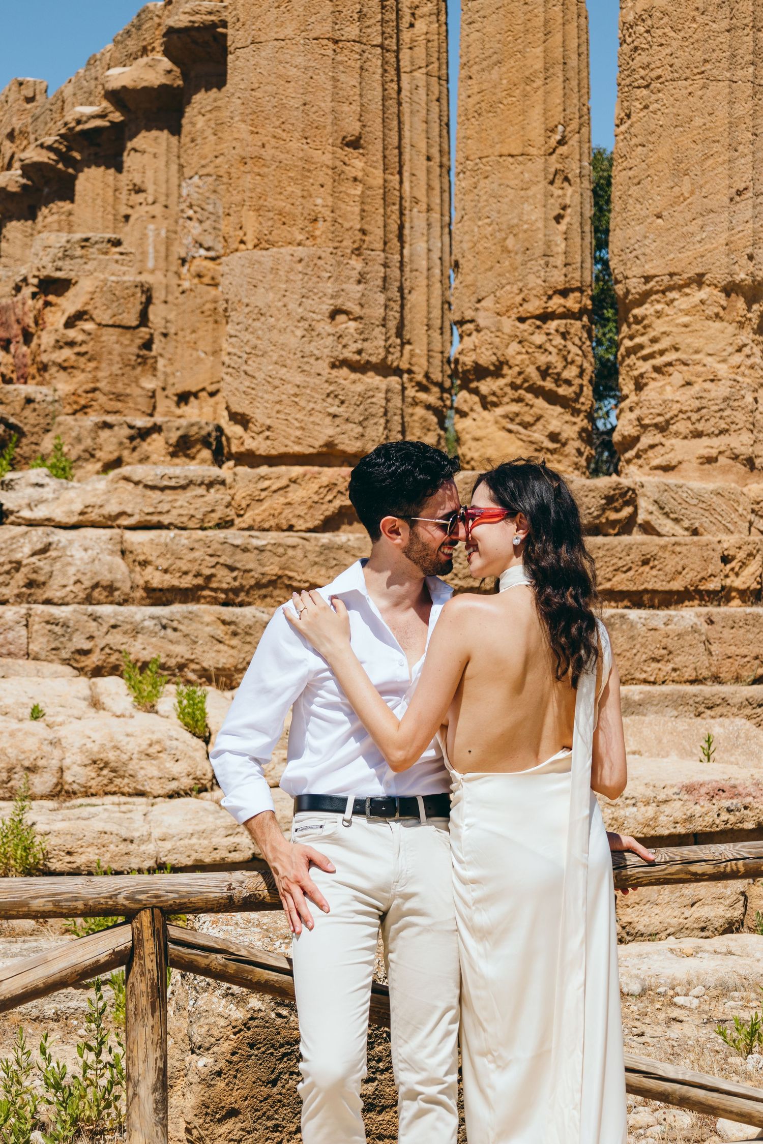 Romantic photoshoot with couple in white attire against ancient temple columns and wooden fence.