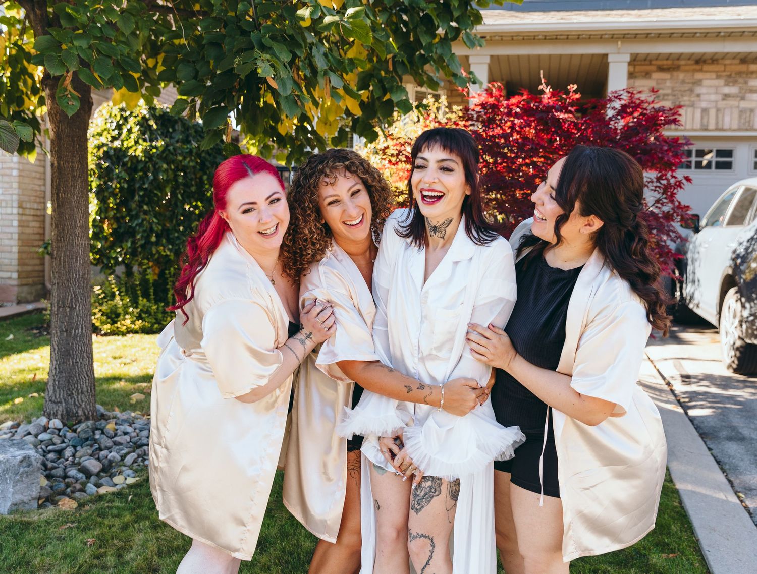 A group in white robes laughs and embraces joyfully outdoors in front of trees and a building.
