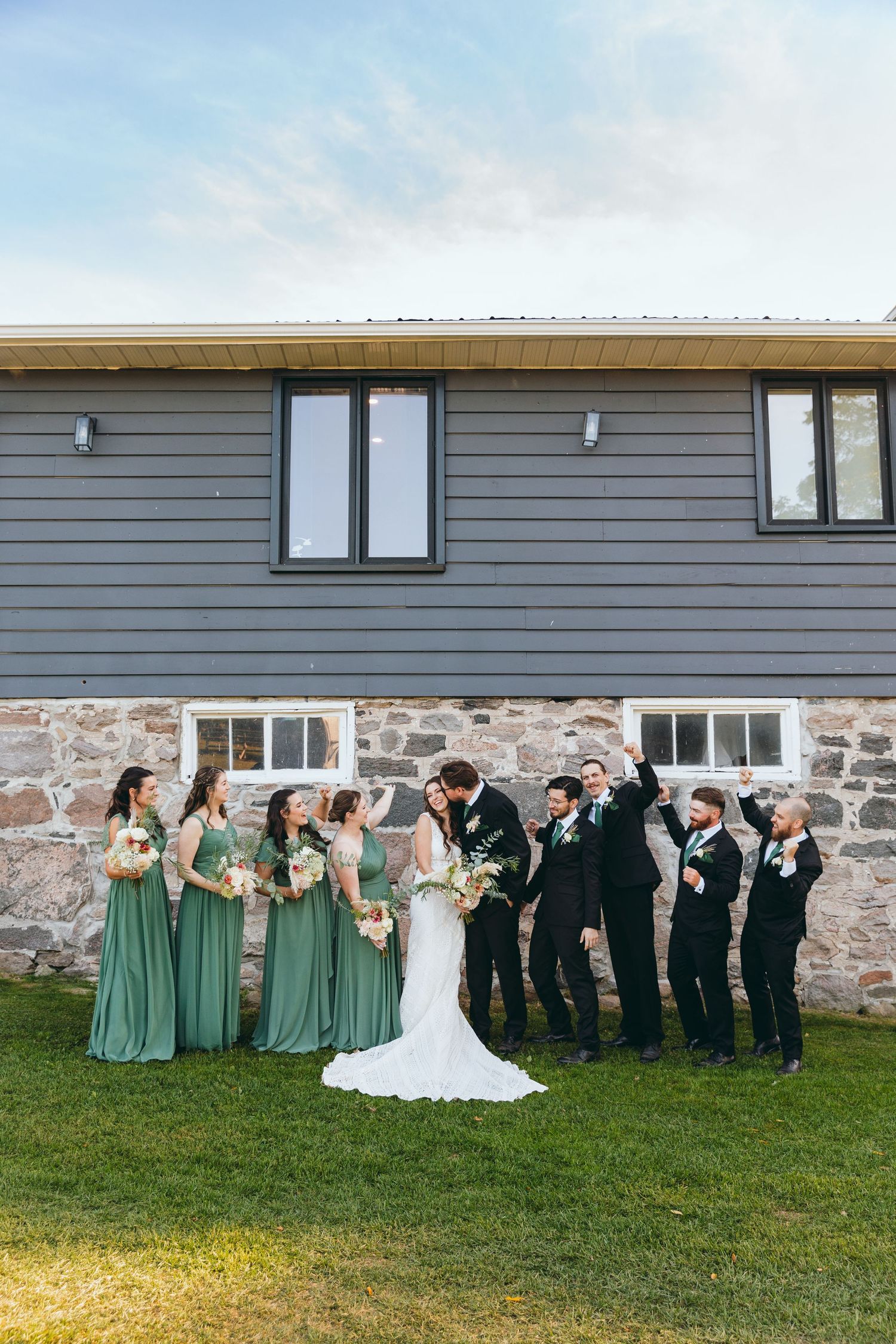 Full wedding party portrait with bridesmaids in green dresses and groomsmen in black suits.