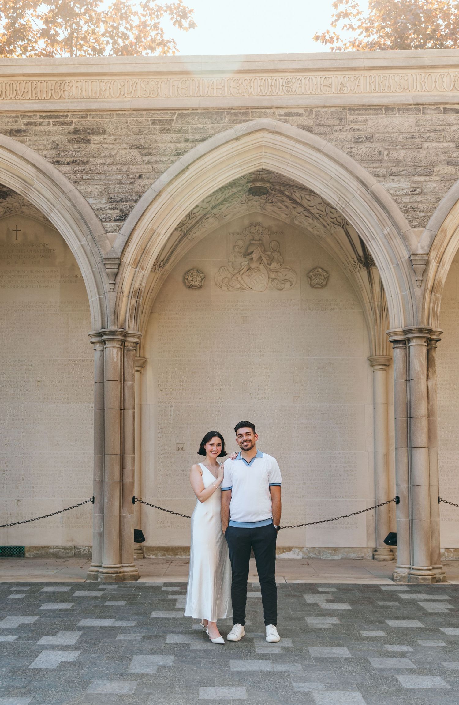 A couple stands together smiling under a gothic stone archway in summer.
