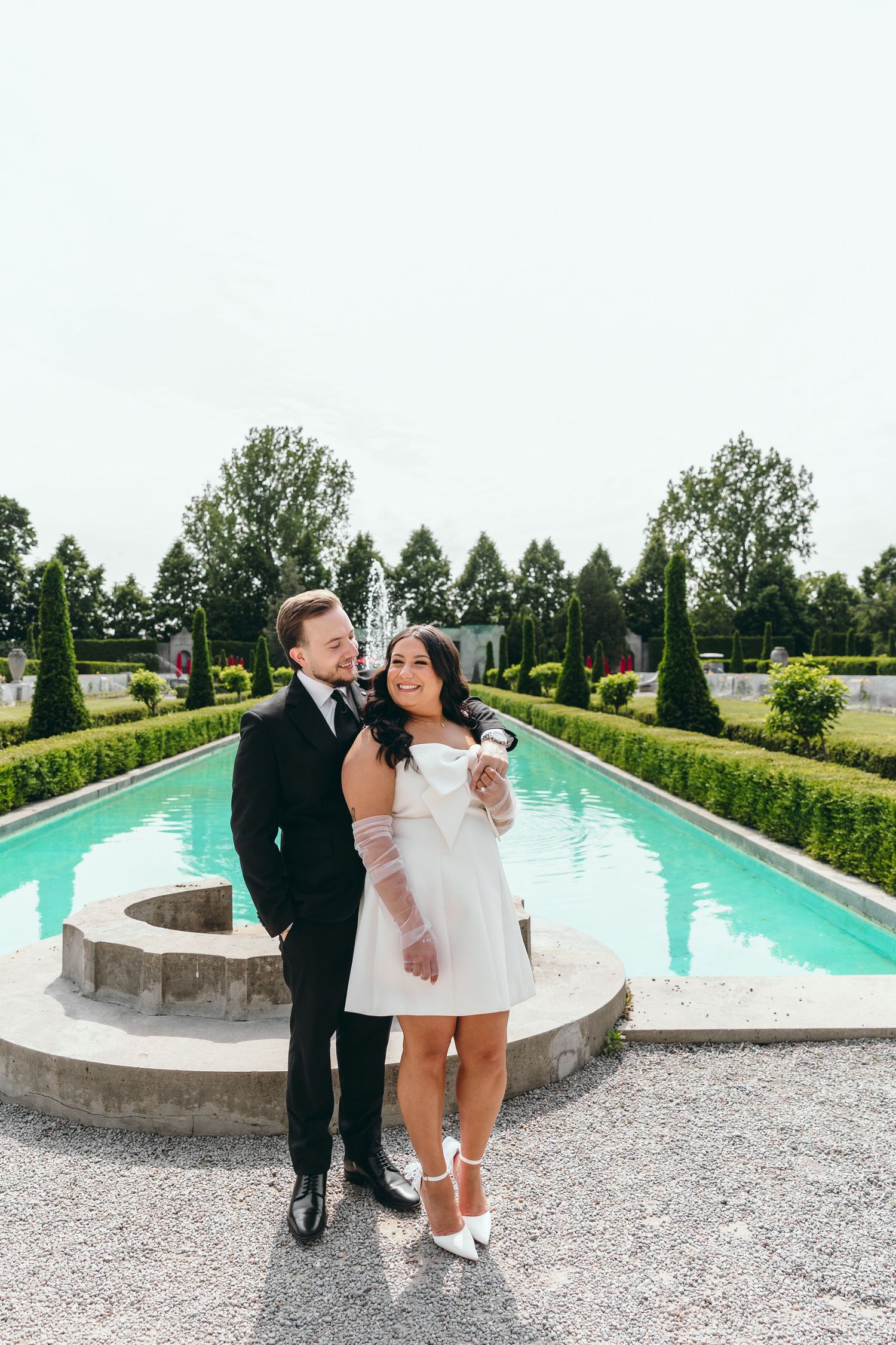 A couple stands together by an elegant pool surrounded by manicured hedges and gardens.