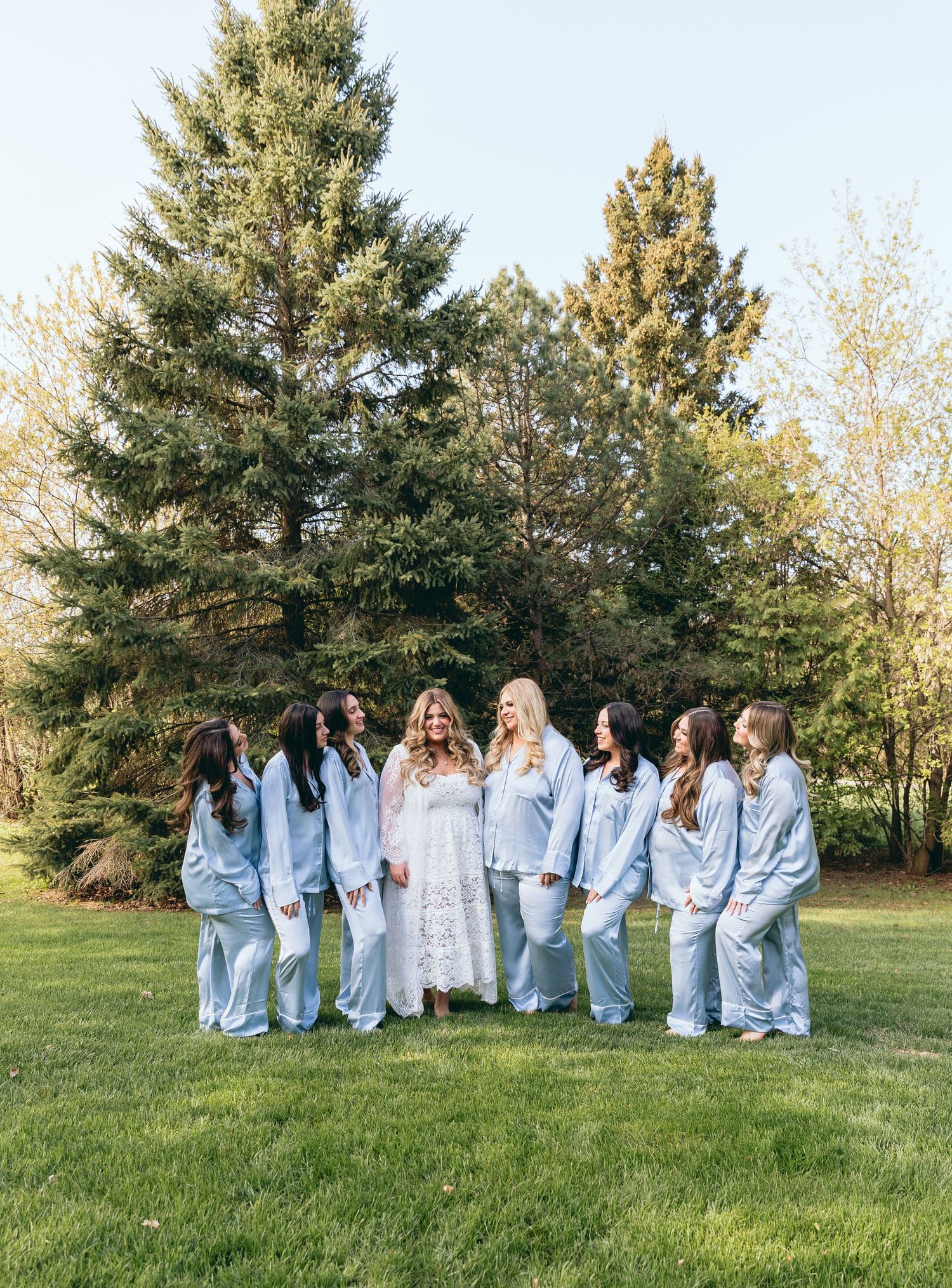 A group wearing light blue robes poses together on a grassy lawn with evergreen trees in the background.