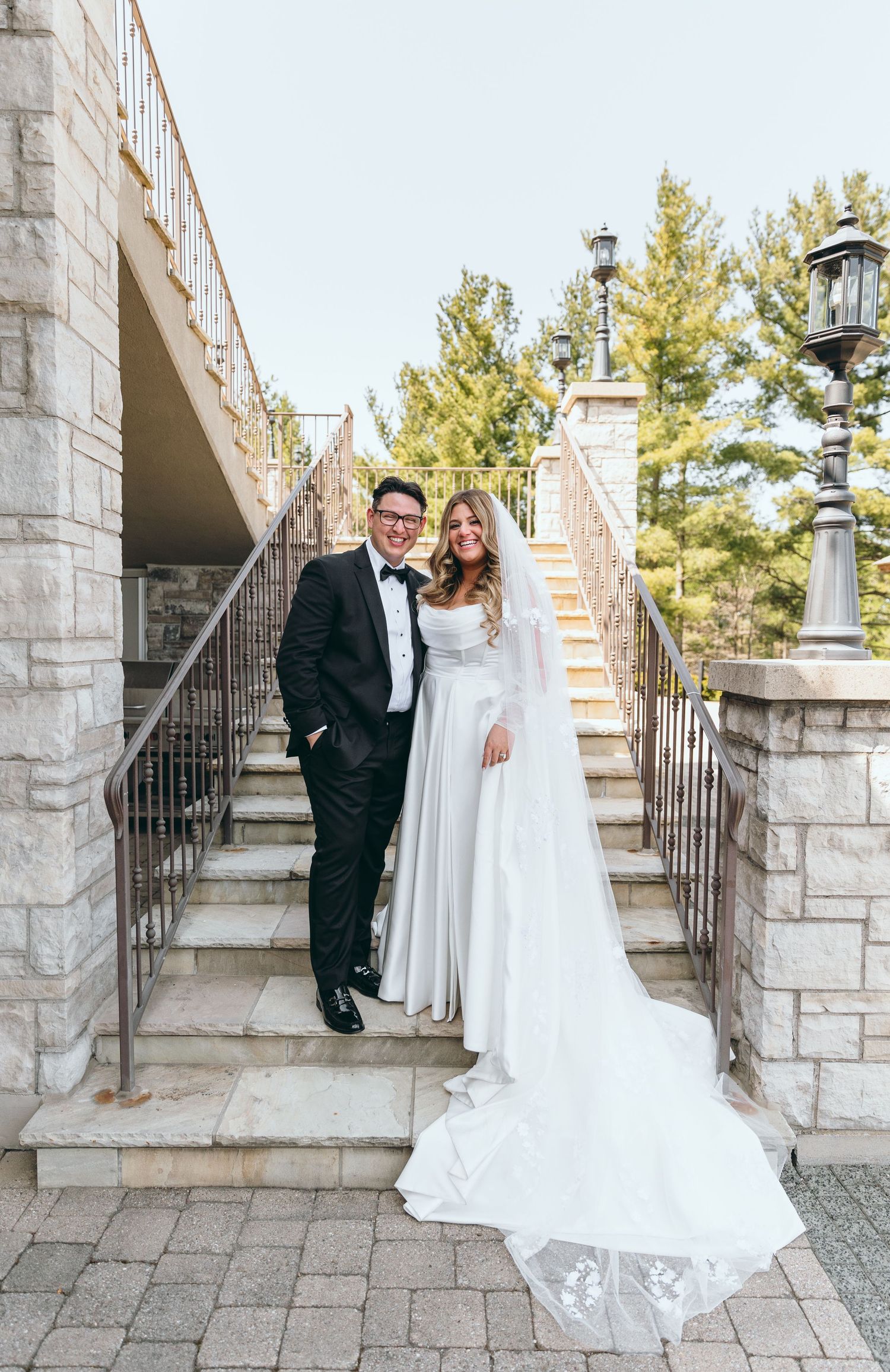 Couple in wedding attire poses on elegant stone staircase with decorative railings.