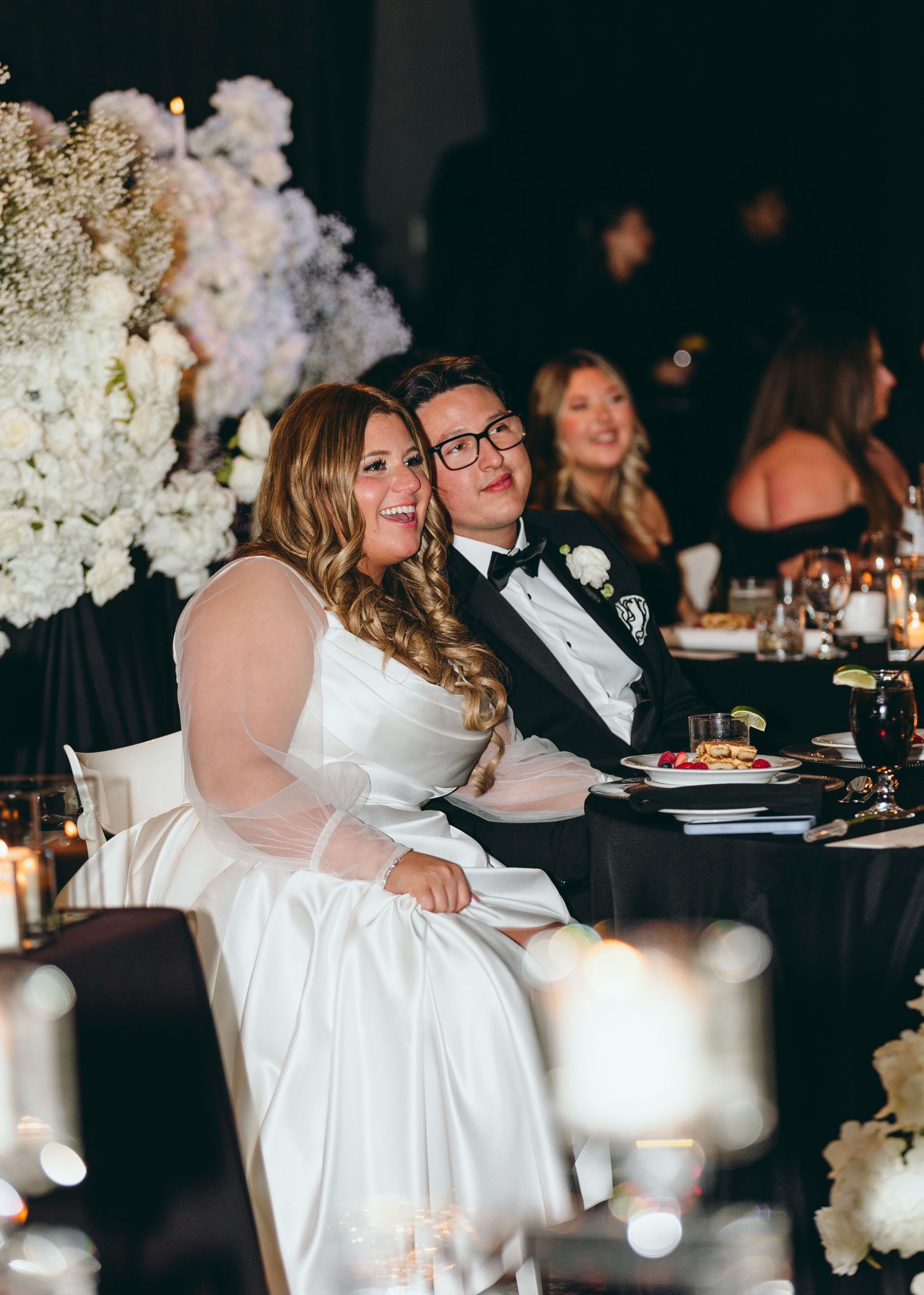 Wedding reception scene with couple at table decorated with white floral arrangements.
