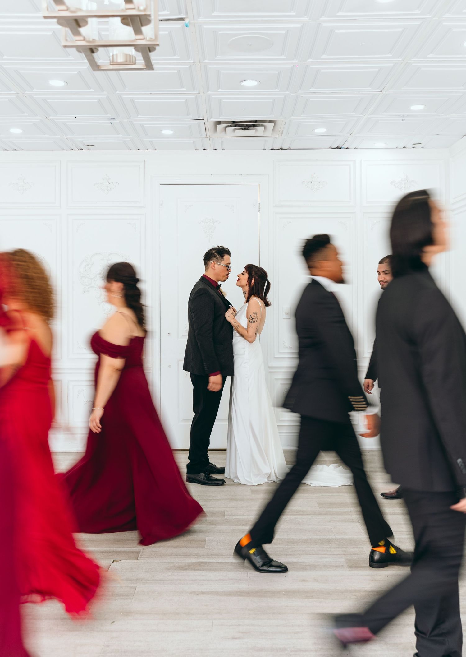 Motion blur captures wedding party dancing in black suits and red dresses against white walls.
