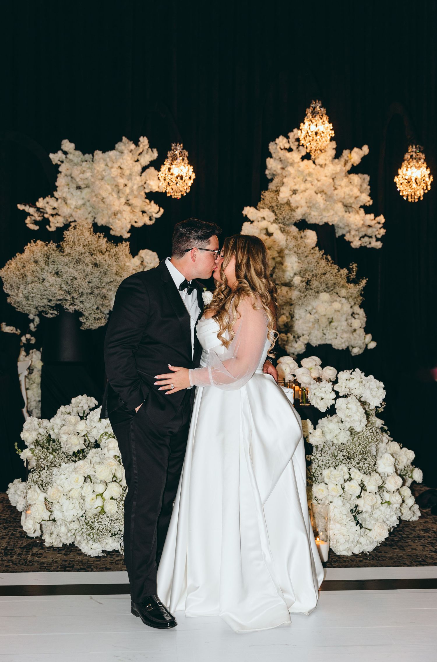 Couple shares romantic moment in front of white floral wall display at wedding reception.