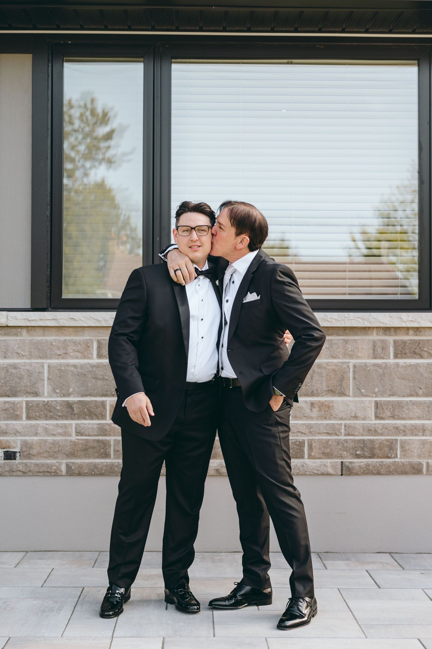 Two people in formal attire sharing a playful moment against a modern building exterior.