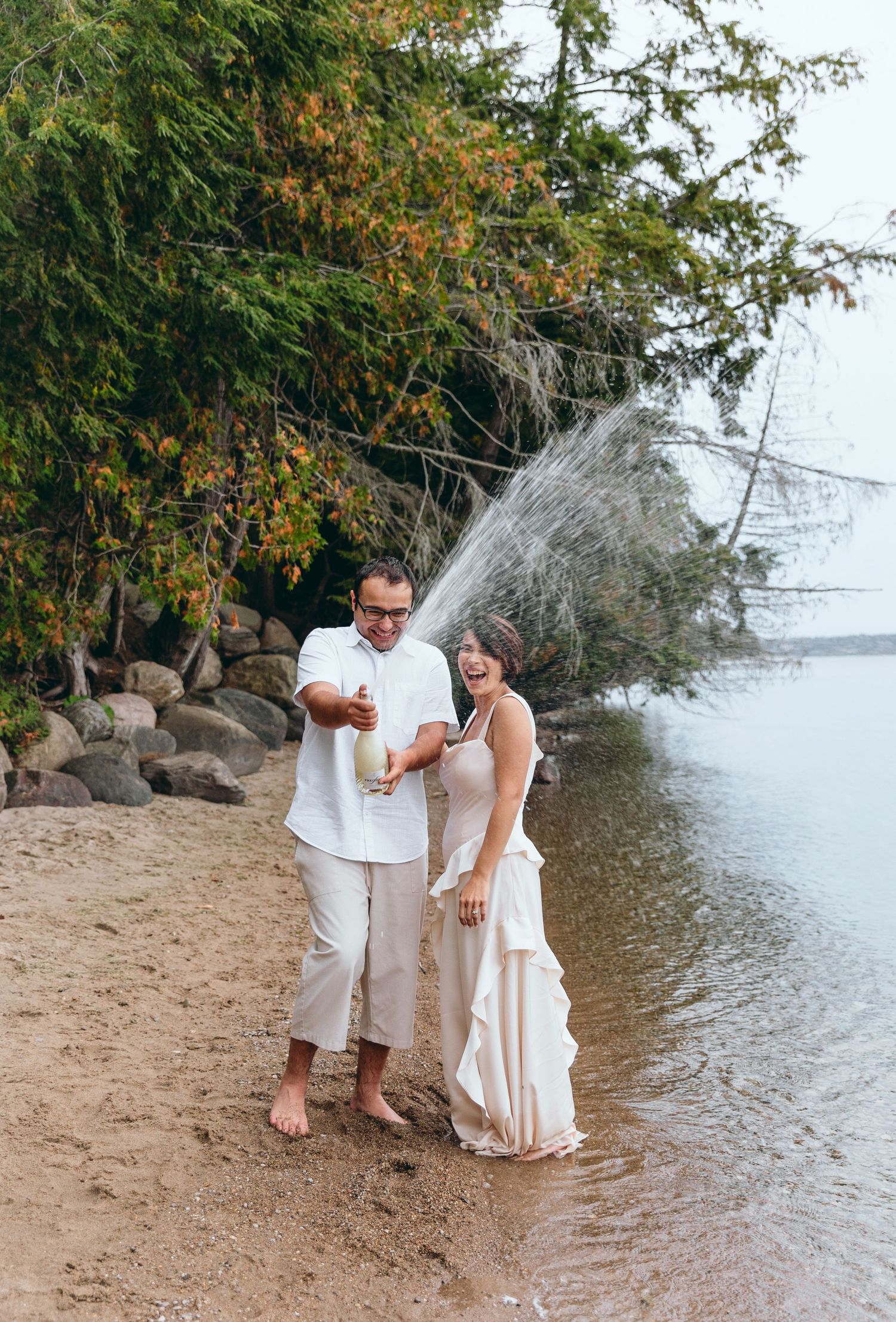 Couple in white clothing splashing water at each other by the lake.