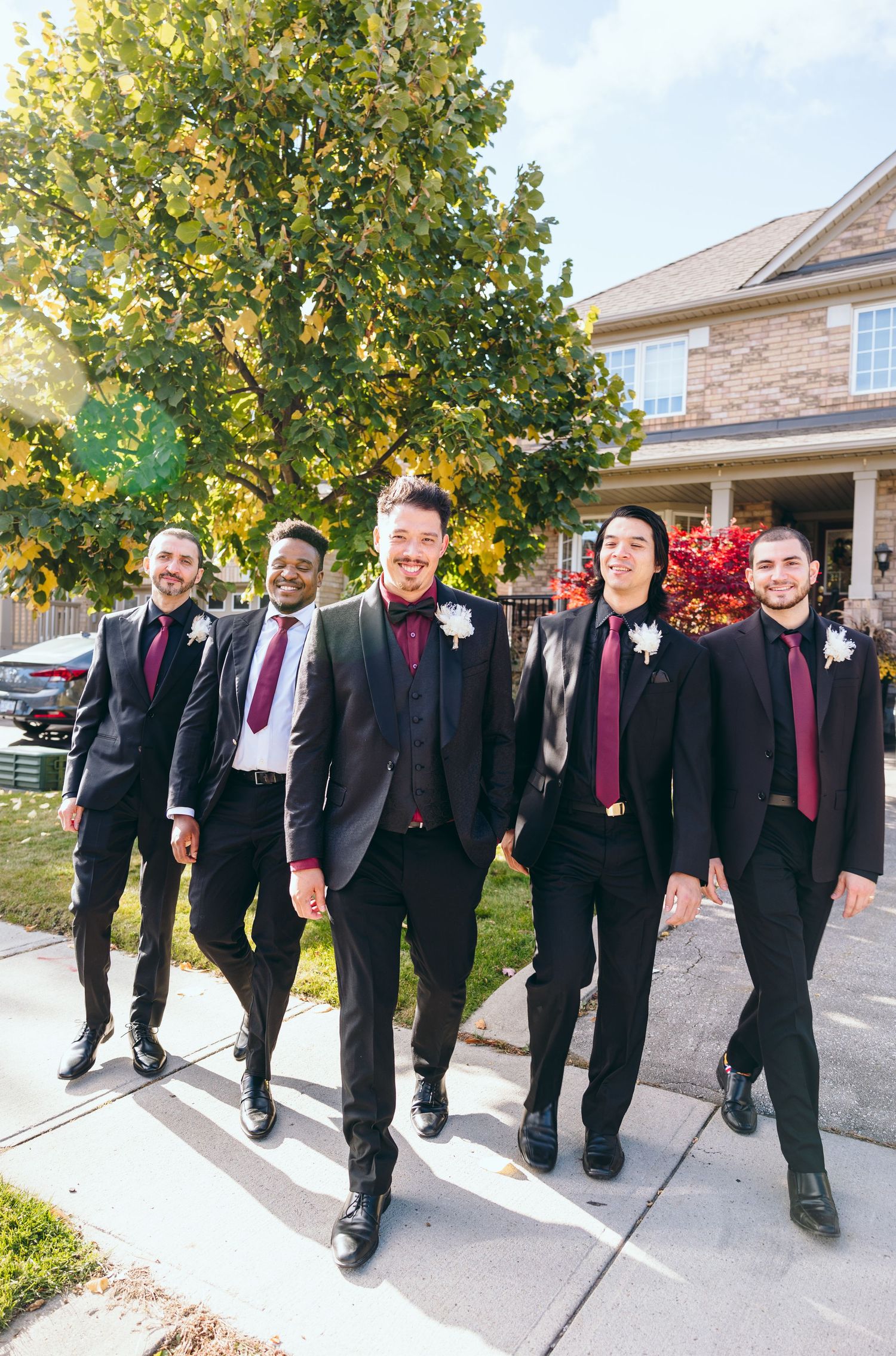 Groomsmen in black suits with burgundy ties walk together on a sidewalk.