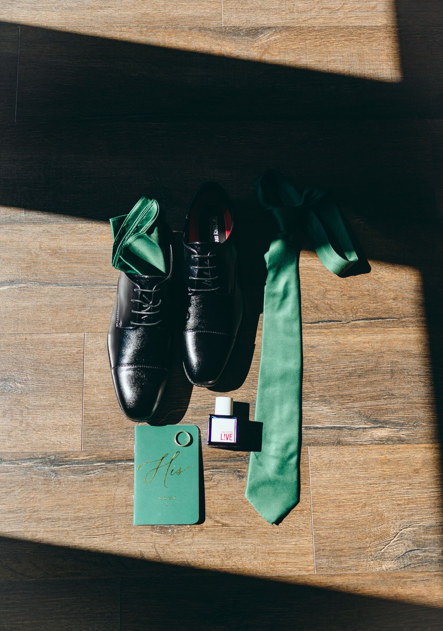 Groom's accessories including black shoes, green tie and stationery on wooden floor.