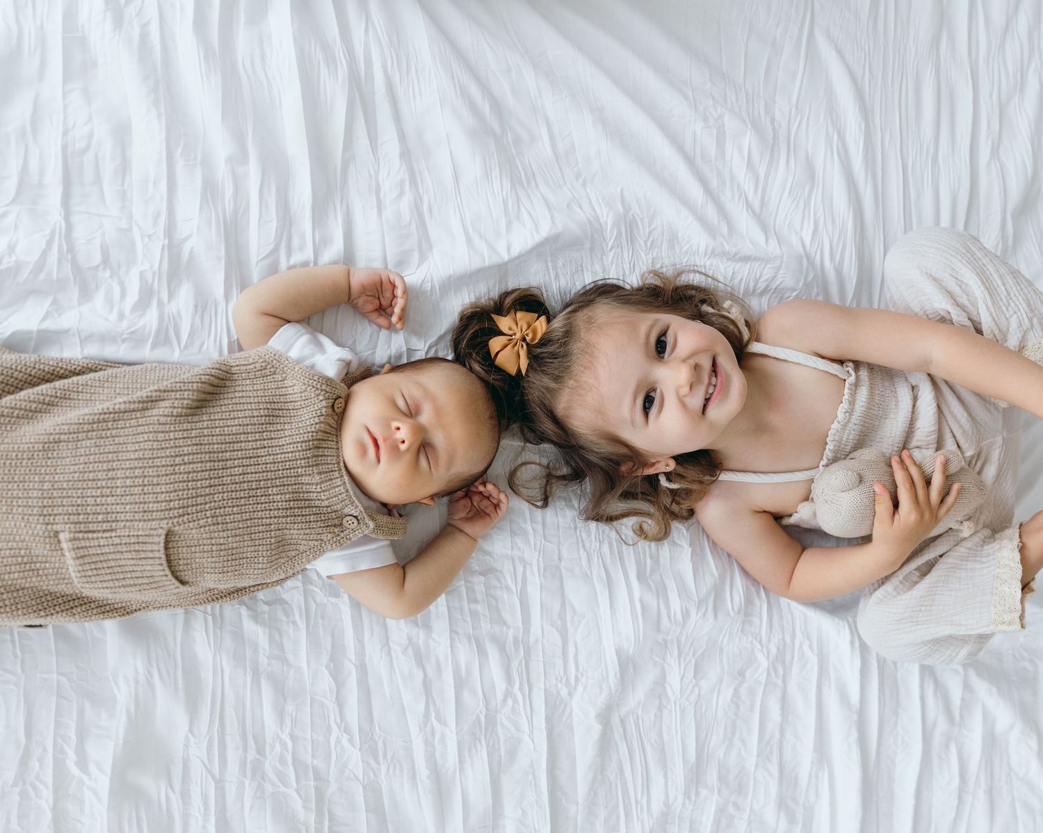 Two children lying on white bedding wearing neutral colored clothing in a bright bedroom setting.