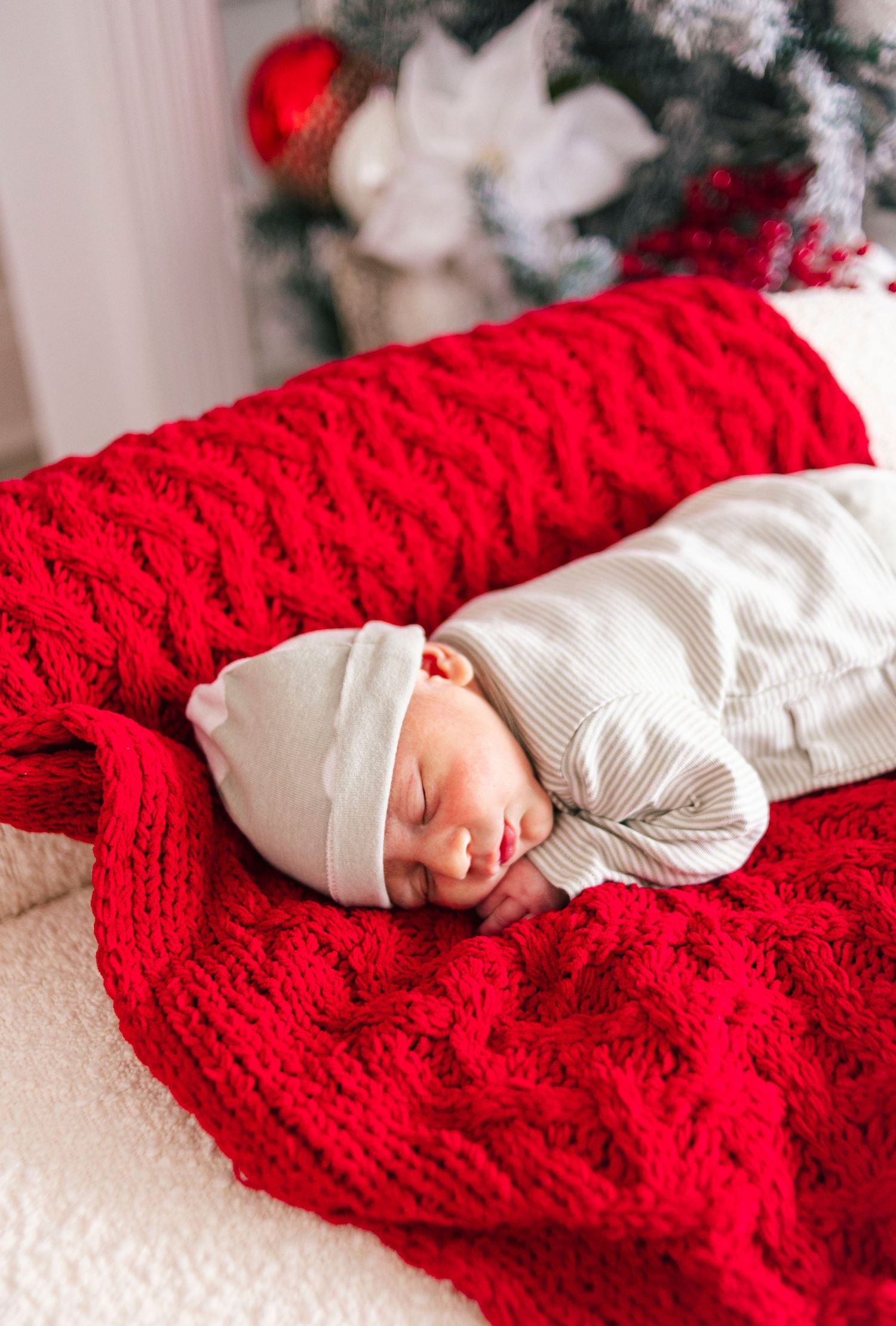 Newborn baby sleeping peacefully on a red knit blanket near a Christmas tree wearing a white striped outfit and hat.