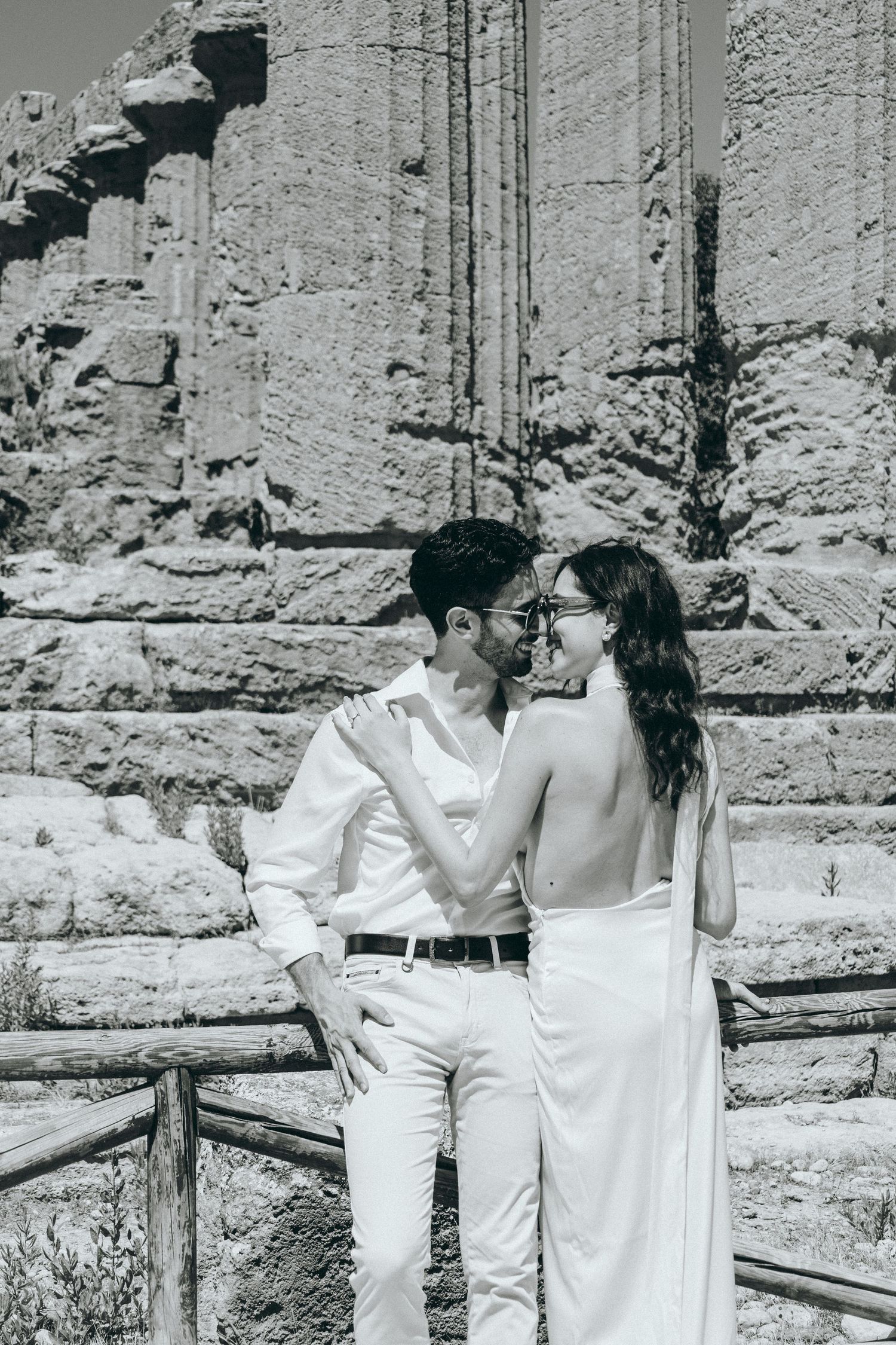 Couple in white outfits pose romantically among ancient Greek ruins.