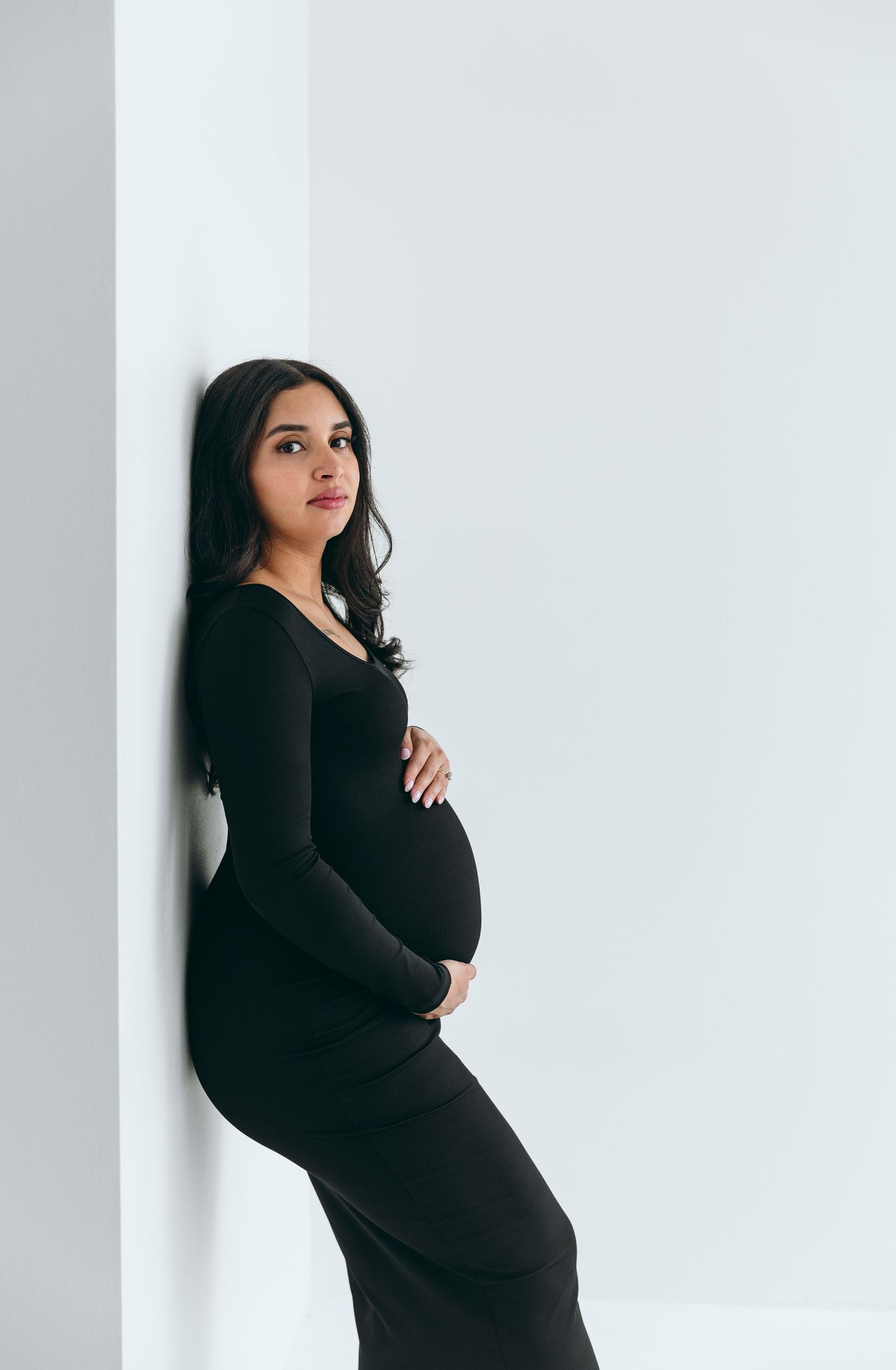Elegant maternity portraits of woman in black dress against white wall.