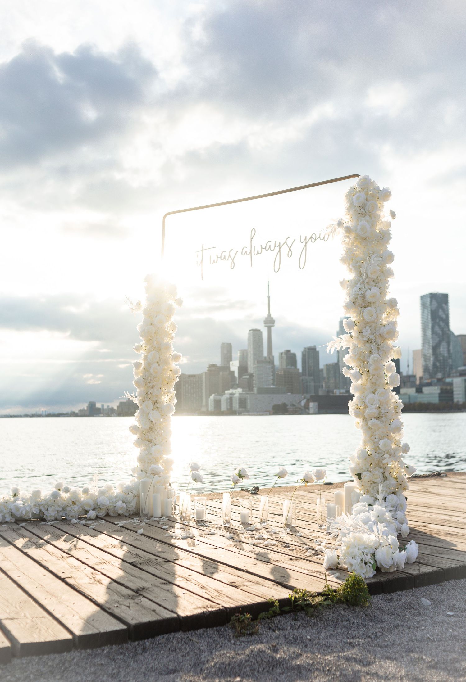 White floral arch setup on wooden deck with city skyline backdrop.