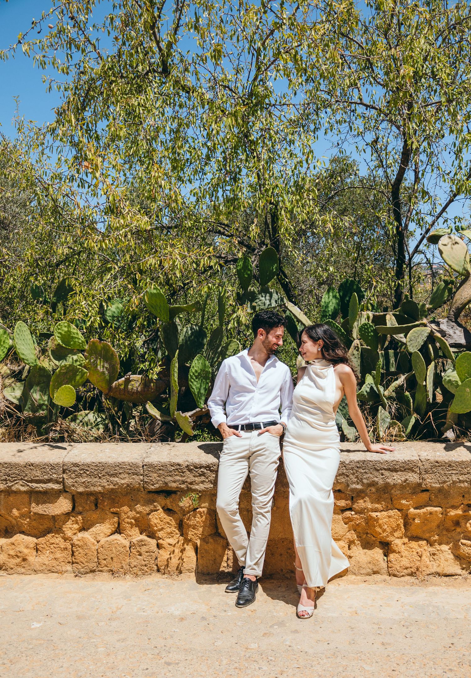 A playful couple relaxes on a stone wall with green trees and cacti in the background.
