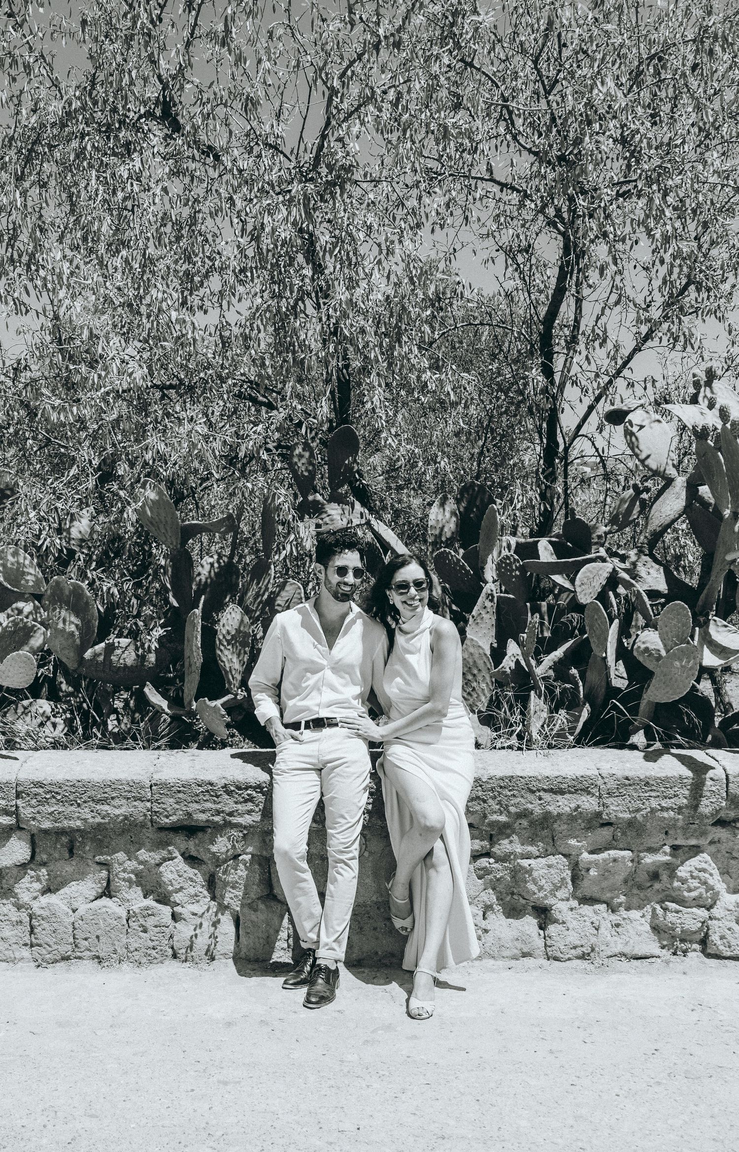 Two people in coordinated white outfits sit casually on a stone wall surrounded by olive trees.
