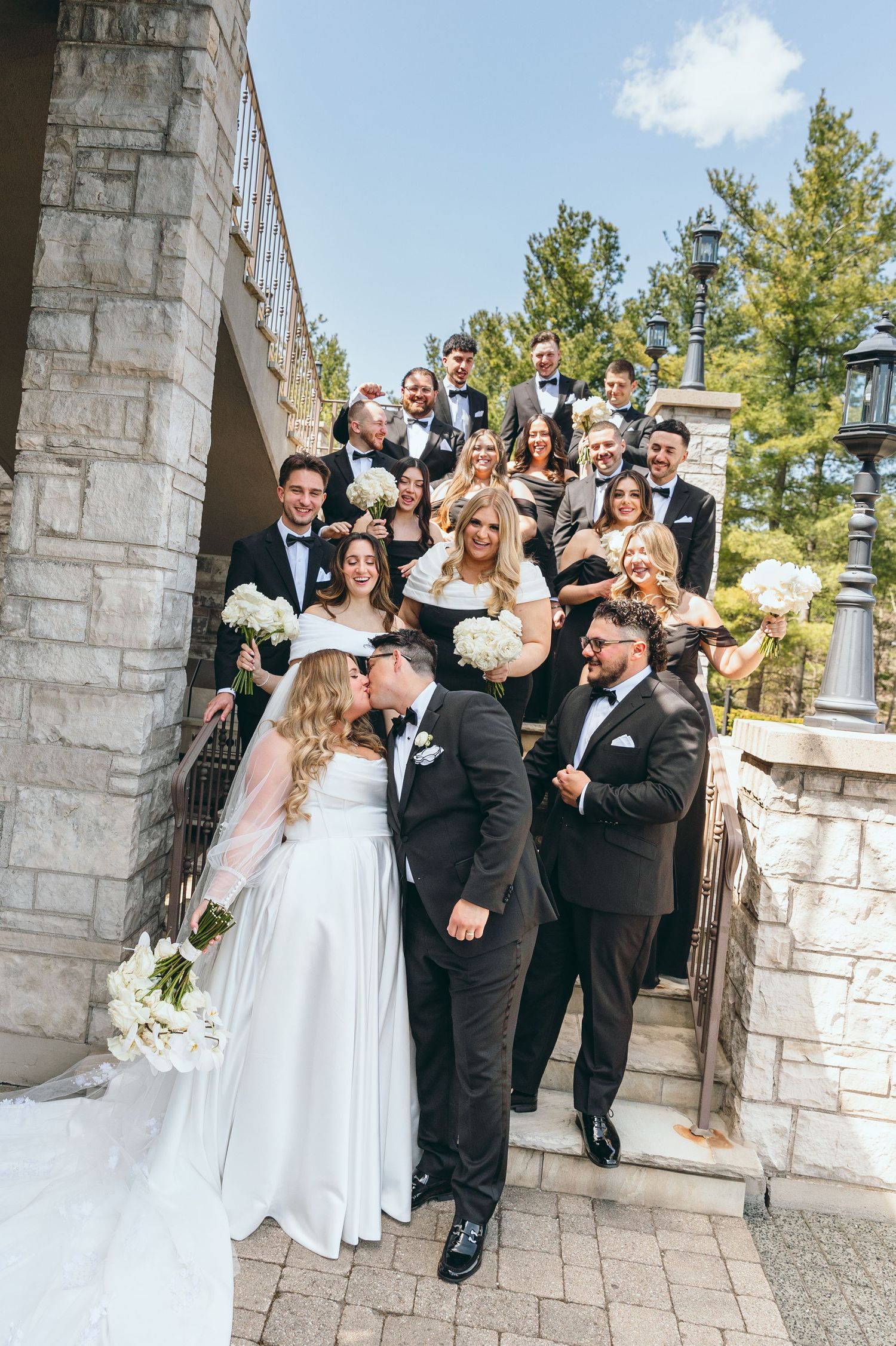 Wedding party poses joyfully on stone steps outside venue in formal attire.