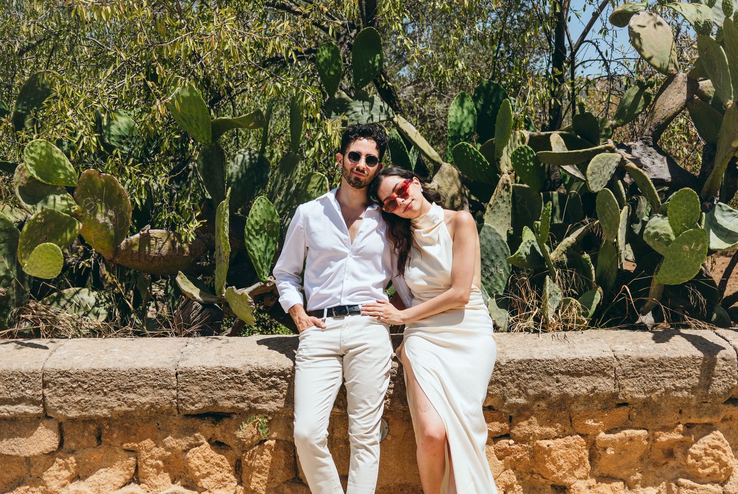 Couple relaxing on stone wall surrounded by desert plants and trees in Mediterranean setting.